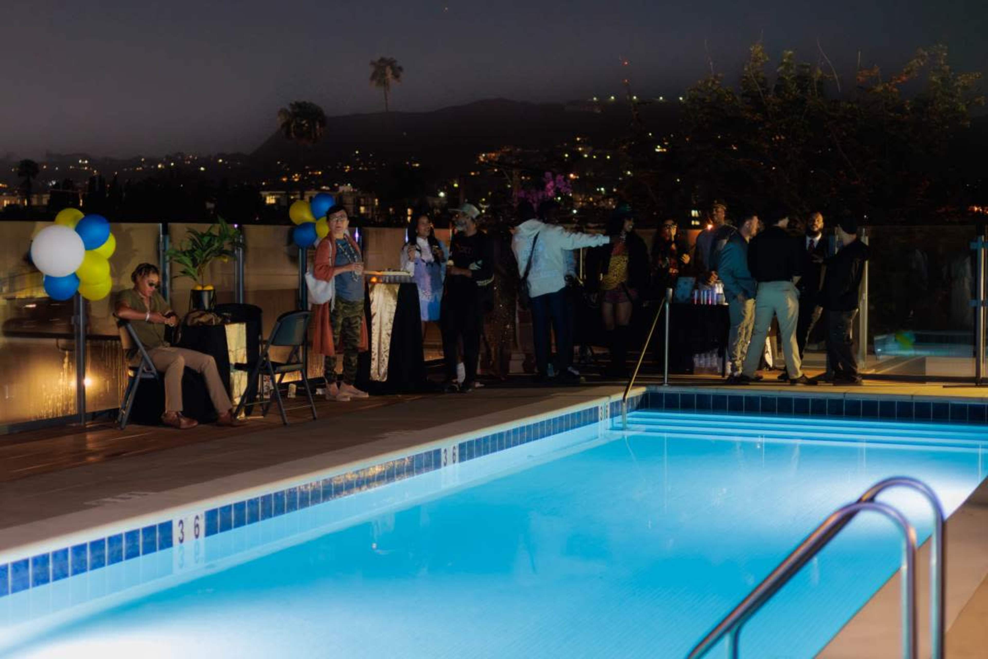 A group of people socializes near a brightly lit pool at an evening event, with a city skyline visible in the background.