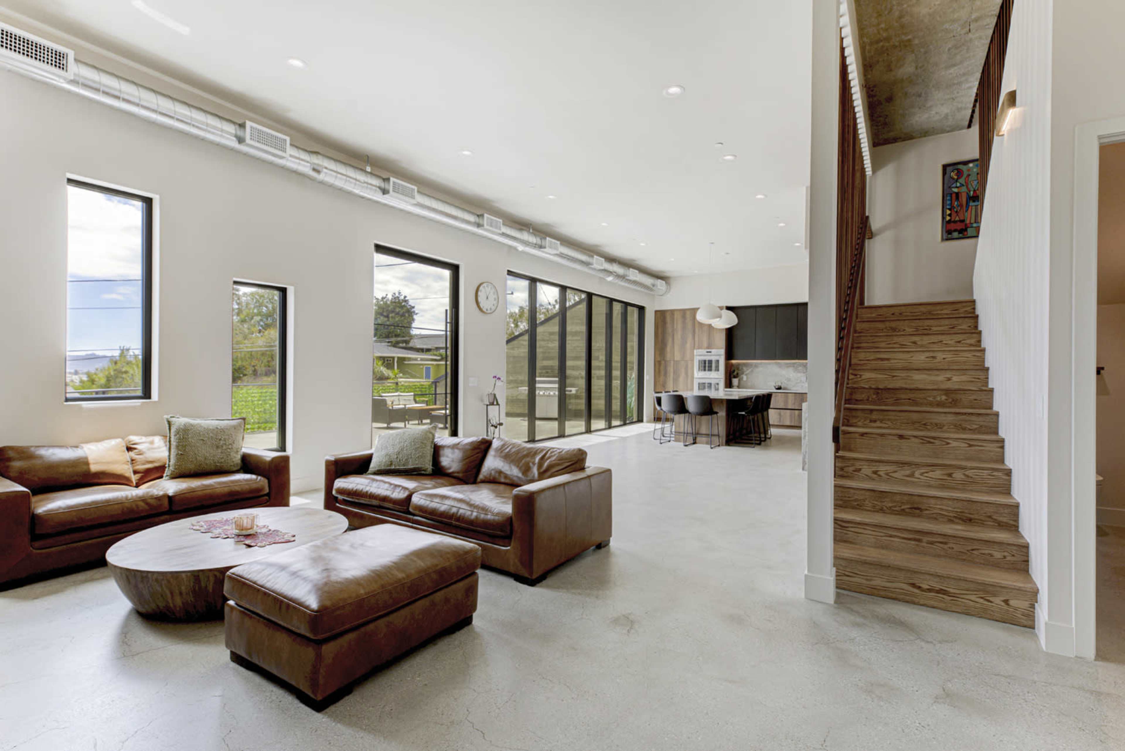 The image shows a spacious, modern living room featuring leather sofas, large windows allowing natural light, and a staircase leading to an upper level.