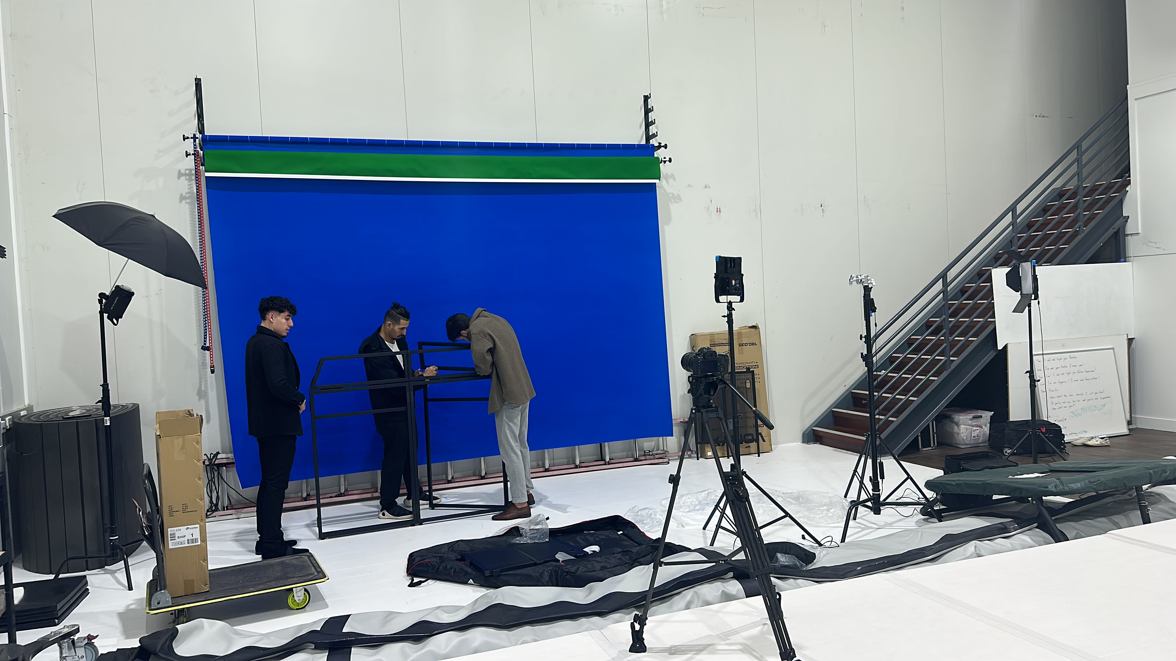 Three individuals in tailored suits are assembling a studio backdrop in a photography studio with a blue screen and various lighting equipment.