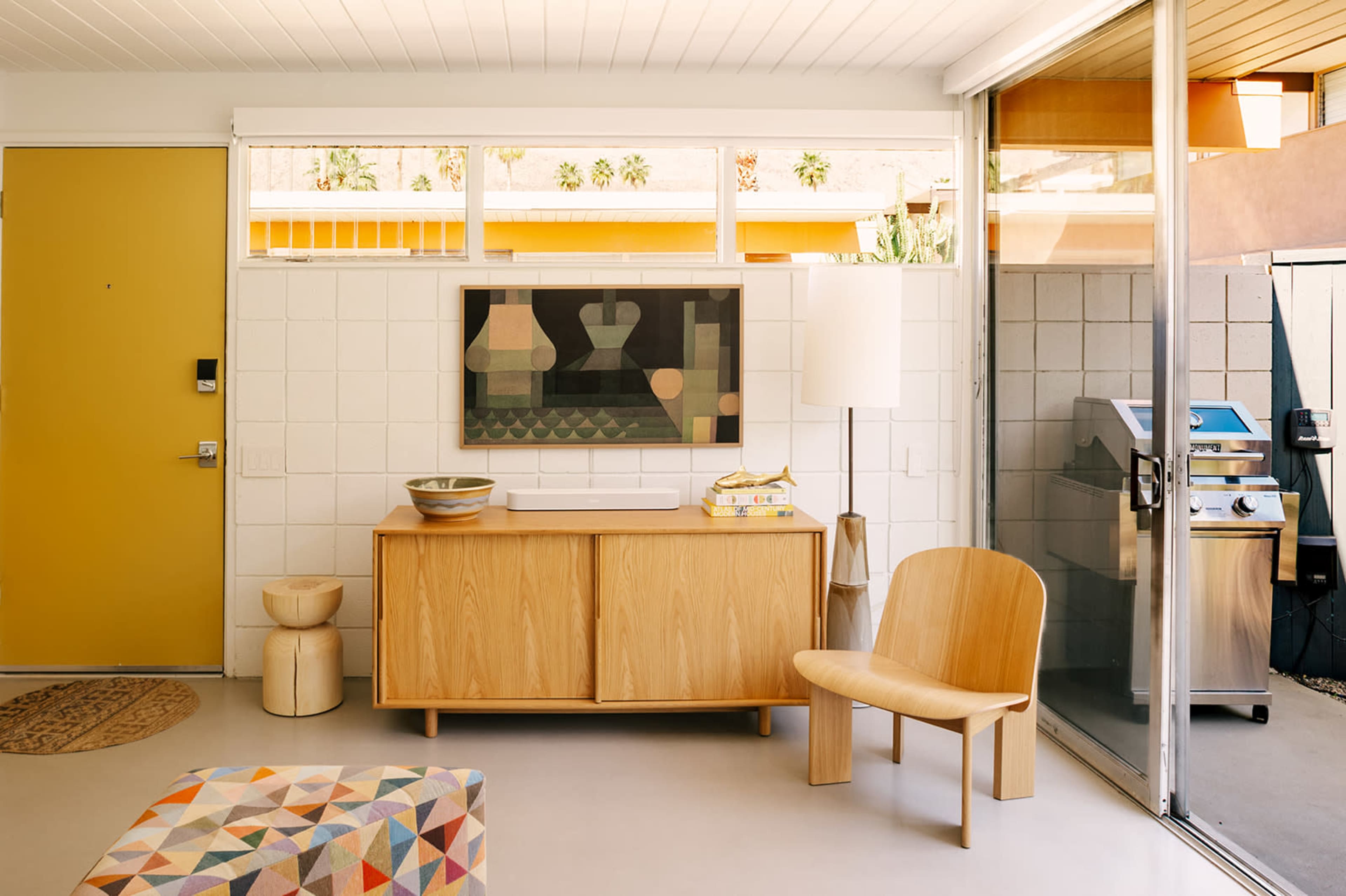 The image shows a modern living room with a yellow front door, wooden furniture, a framed artwork, and a glass door leading to an outdoor area with a grill.