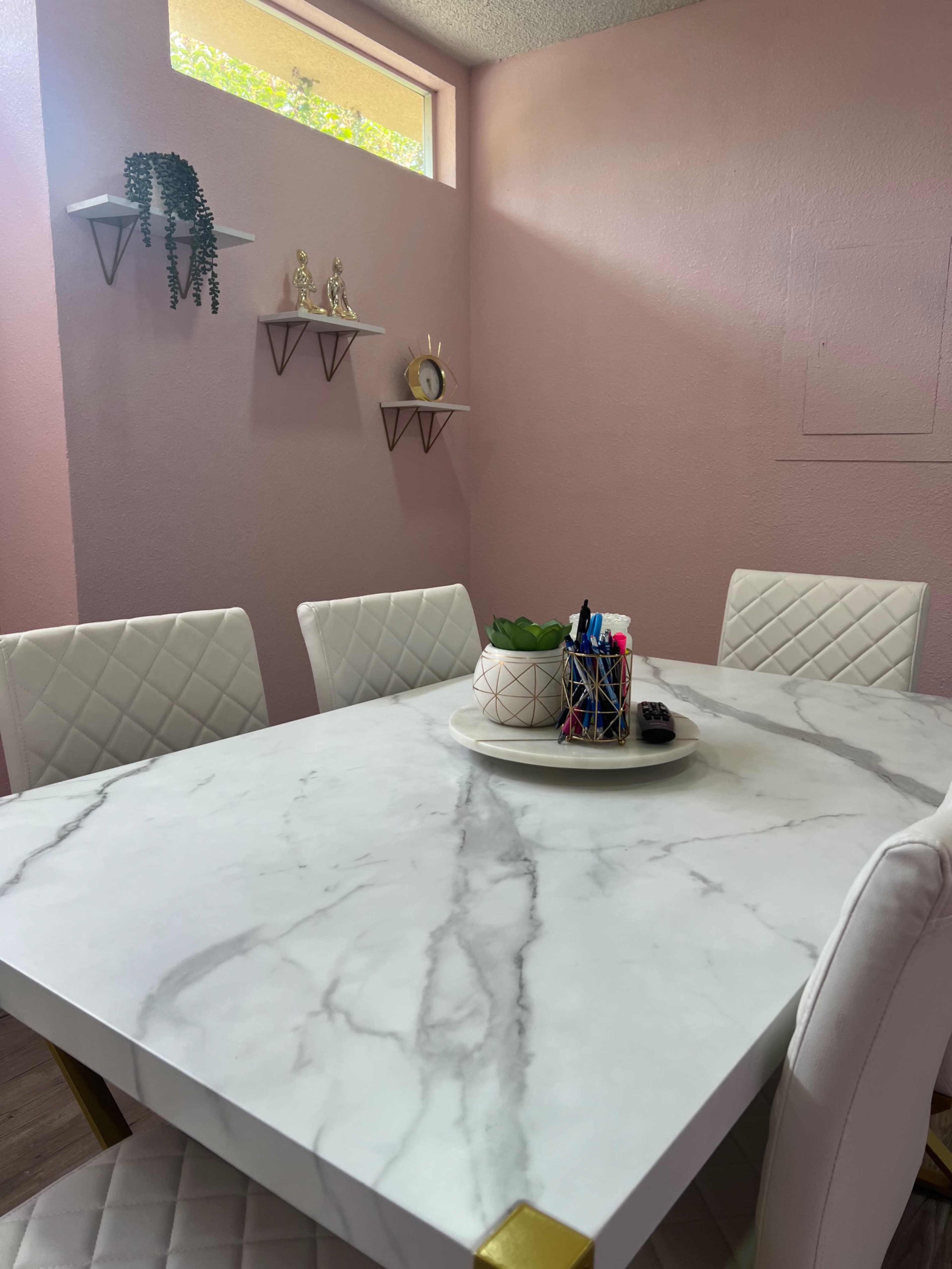 A marble-top table is set in a room with pink walls, surrounded by white, quilted chairs and adorned with decorative shelves and a plant.