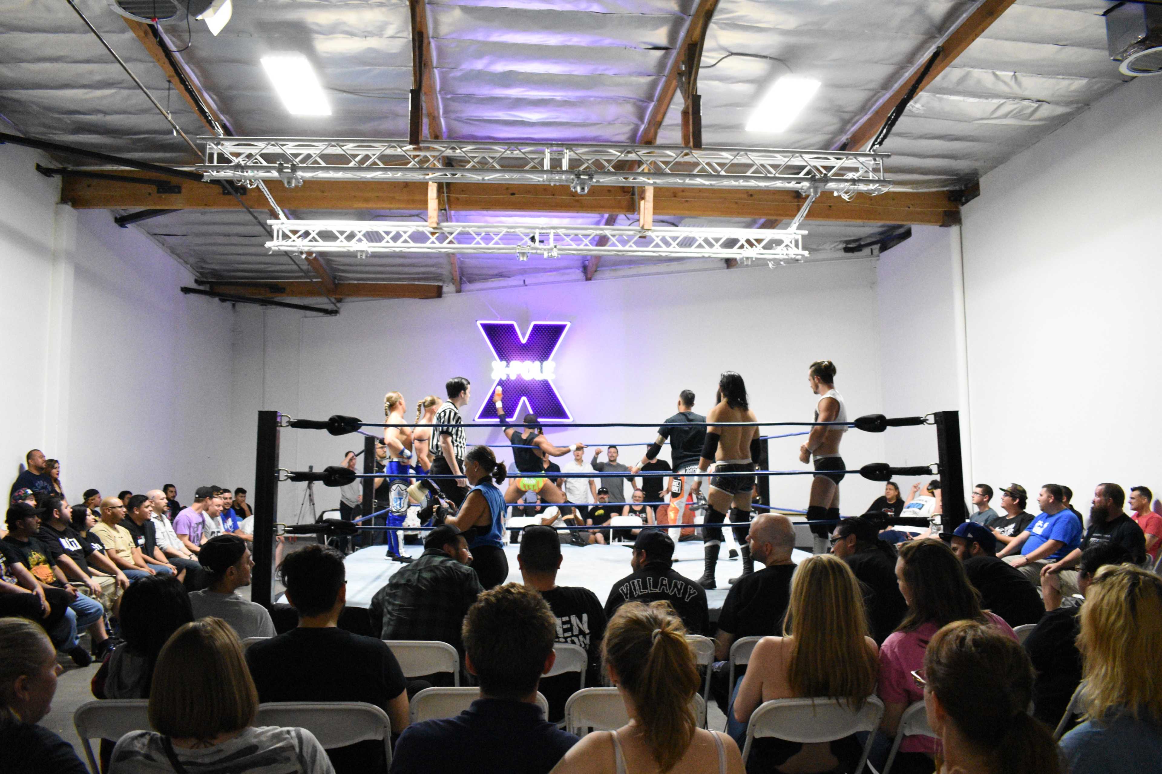 A wrestling event takes place in an indoor arena with a ring surrounded by spectators seated in folding chairs.