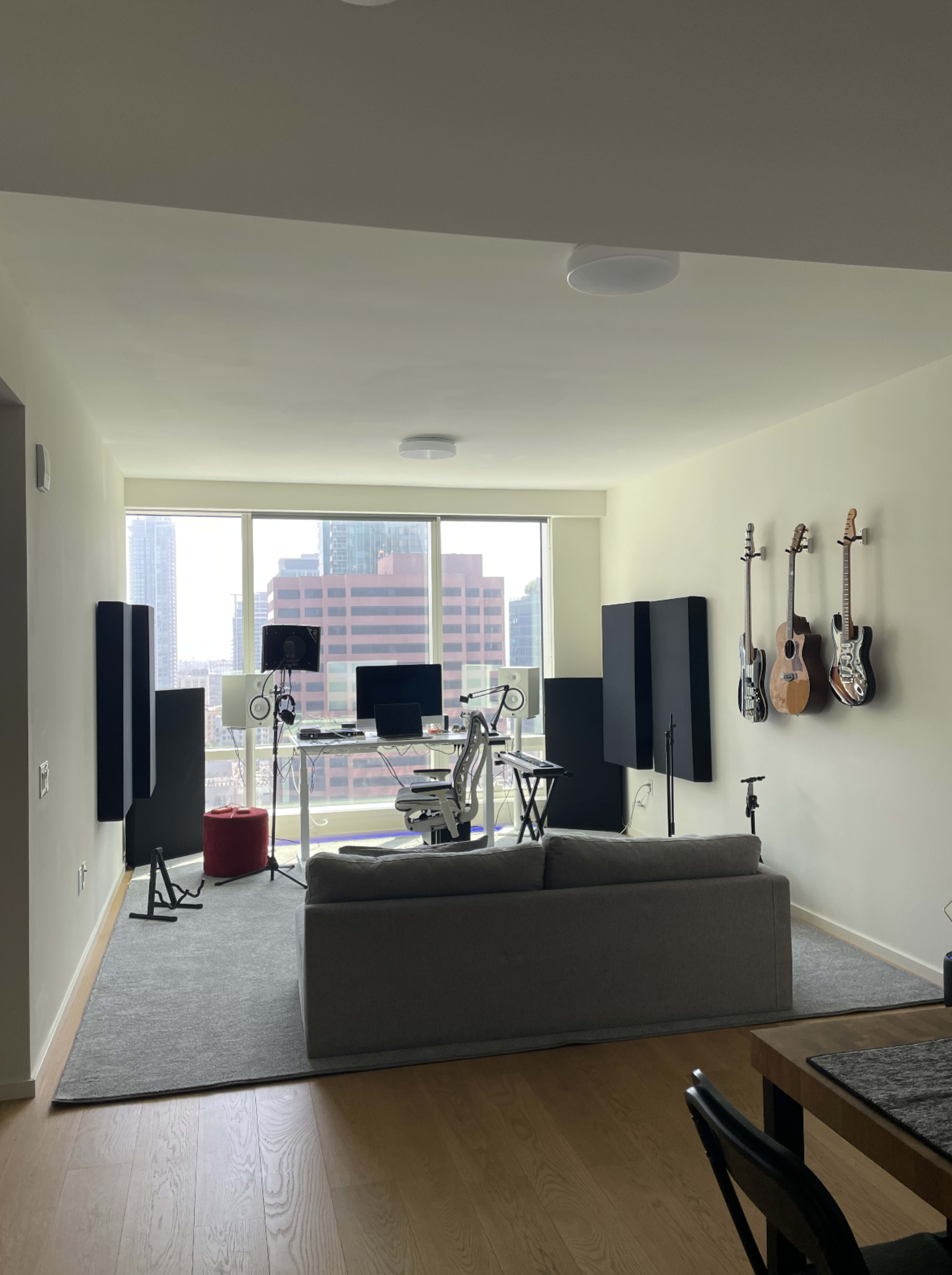 A modern living space features a sofa, a workstation with monitors and audio equipment, and guitars mounted on the wall, with large windows providing a view of a city skyline.