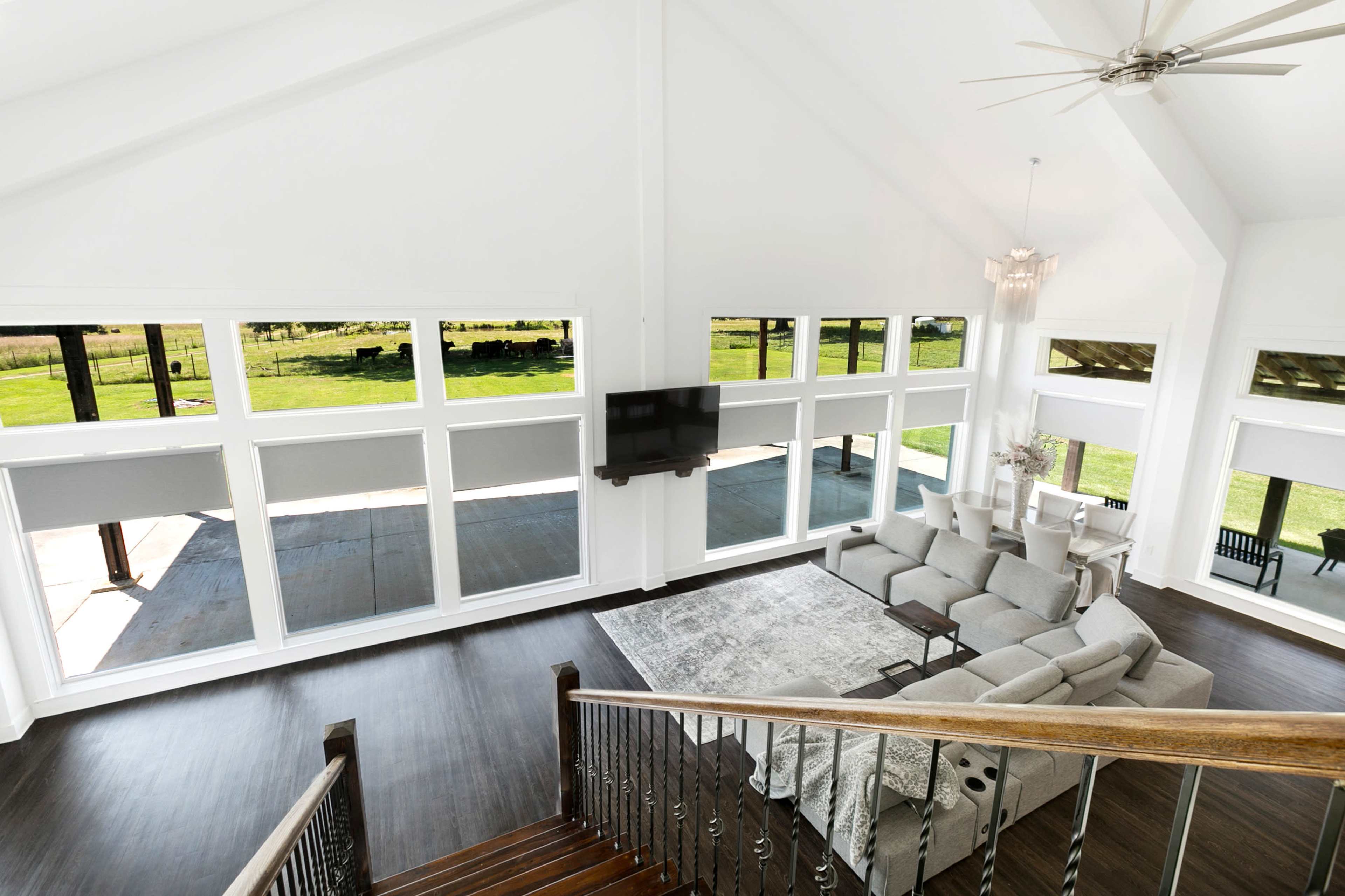 The image shows a spacious, modern living room with large windows, hardwood floors, and a sectional sofa arranged around a coffee table.
