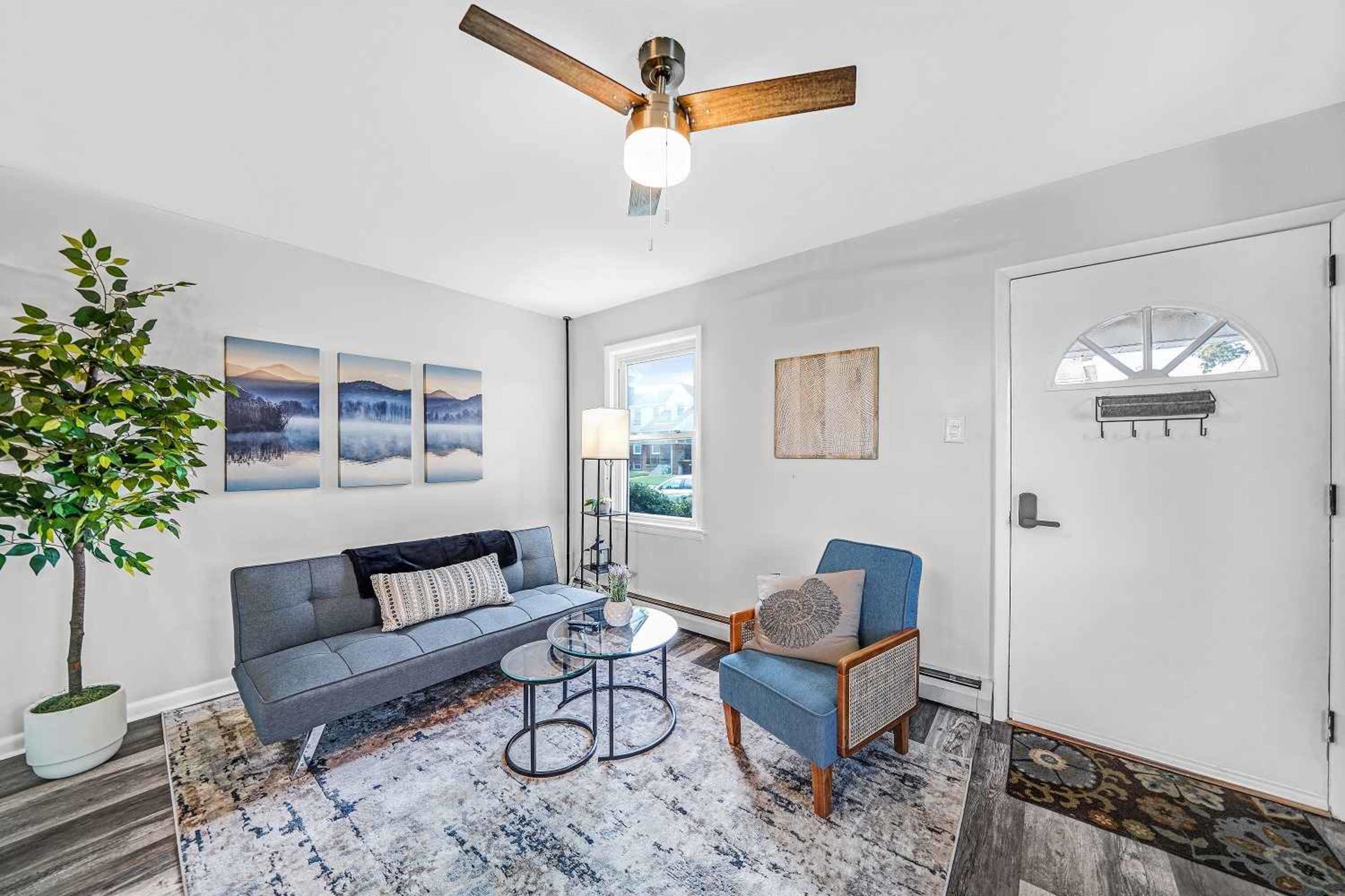 A cozy living room featuring a gray sofa, an armchair, a round table, and a door leading outside, with artwork and a potted plant accenting the space.