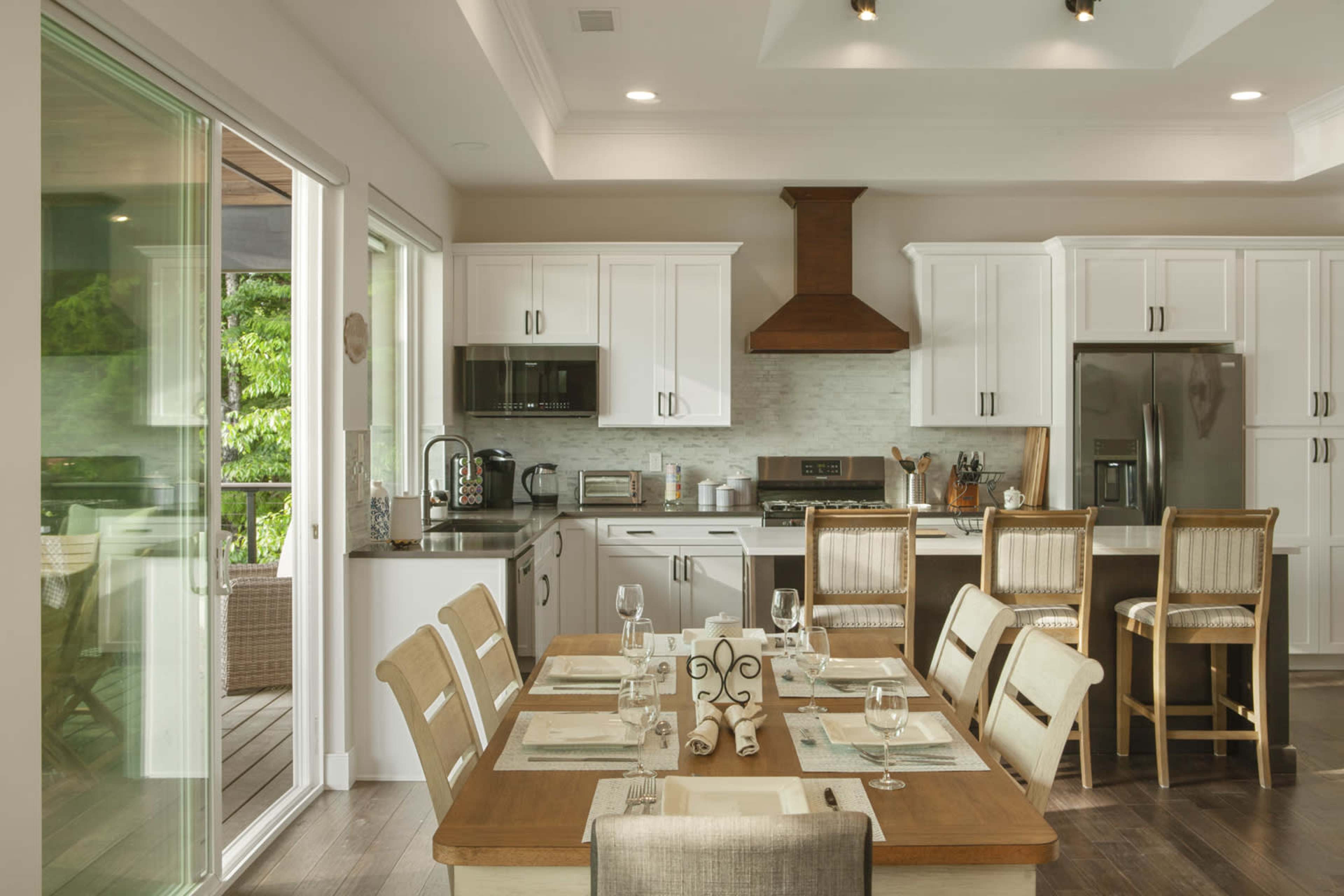 A modern kitchen and dining area features white cabinetry, stainless steel appliances, a wooden dining table set for eight, and a large glass sliding door leading to an outdoor space.