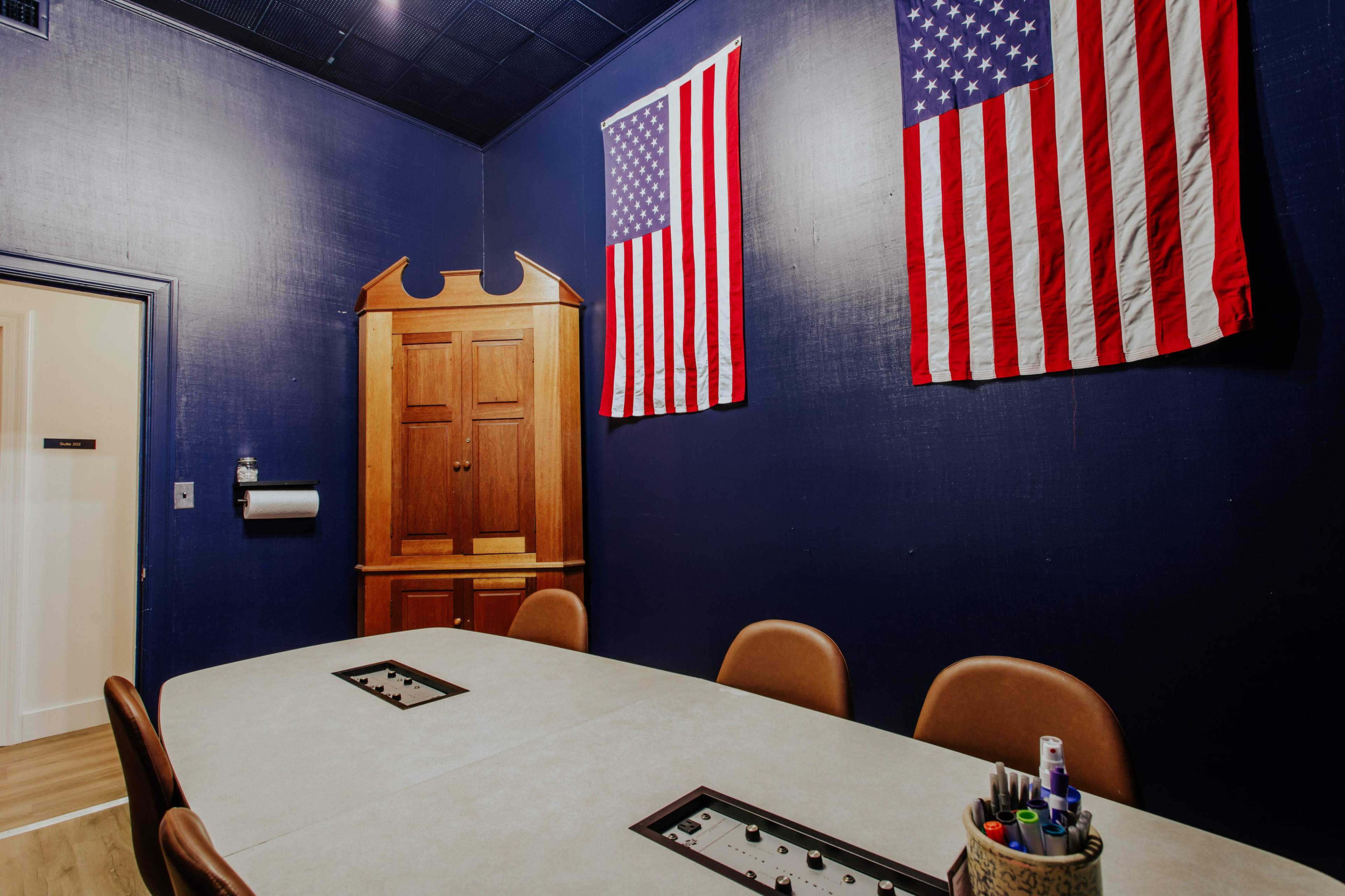 The image shows a conference room with a gray table, brown chairs, and two American flags hanging on the dark blue walls.