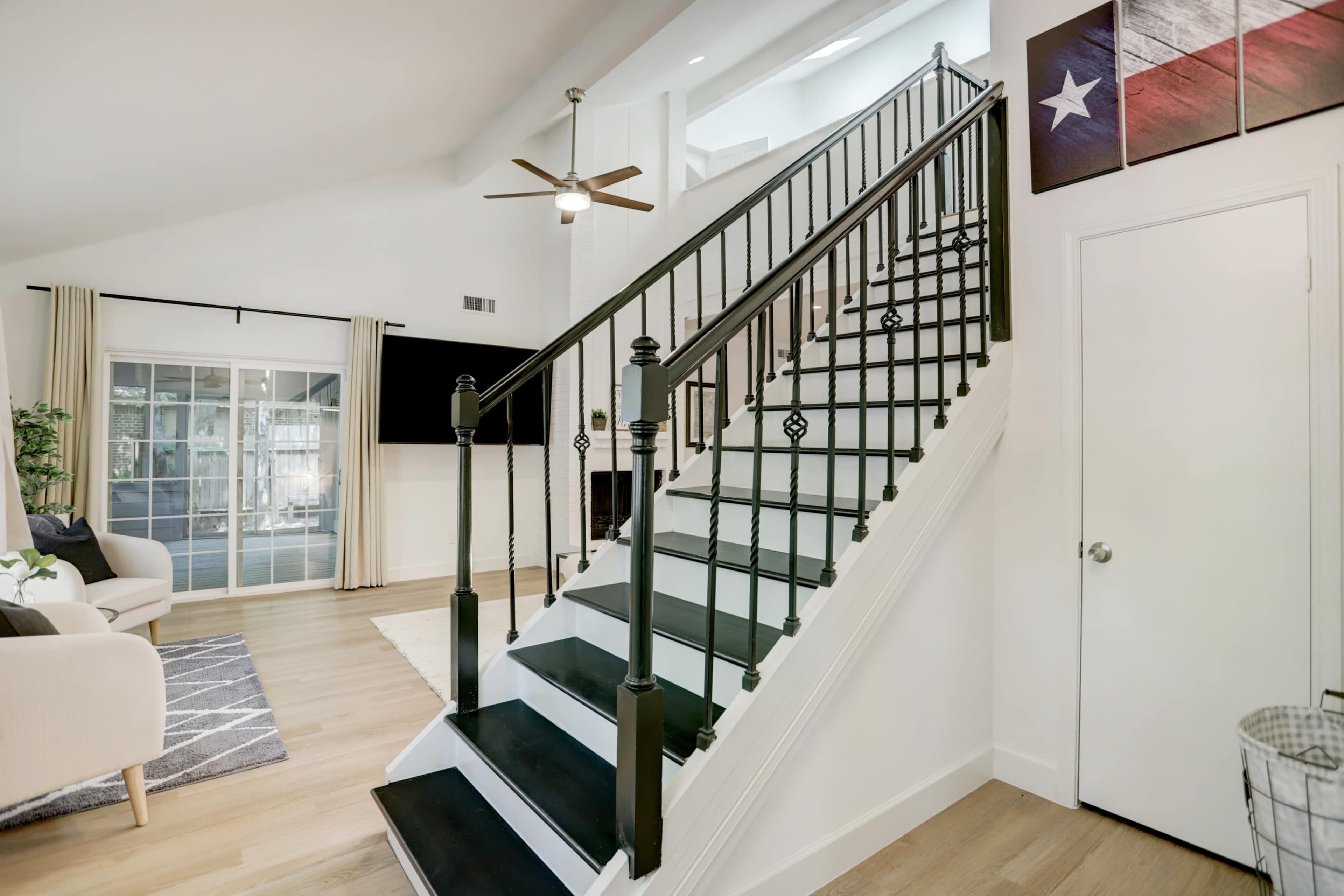 A staircase with black railings leading to an upper level in a well-lit living space, featuring a modern aesthetic and a Texas flag artwork on the wall.