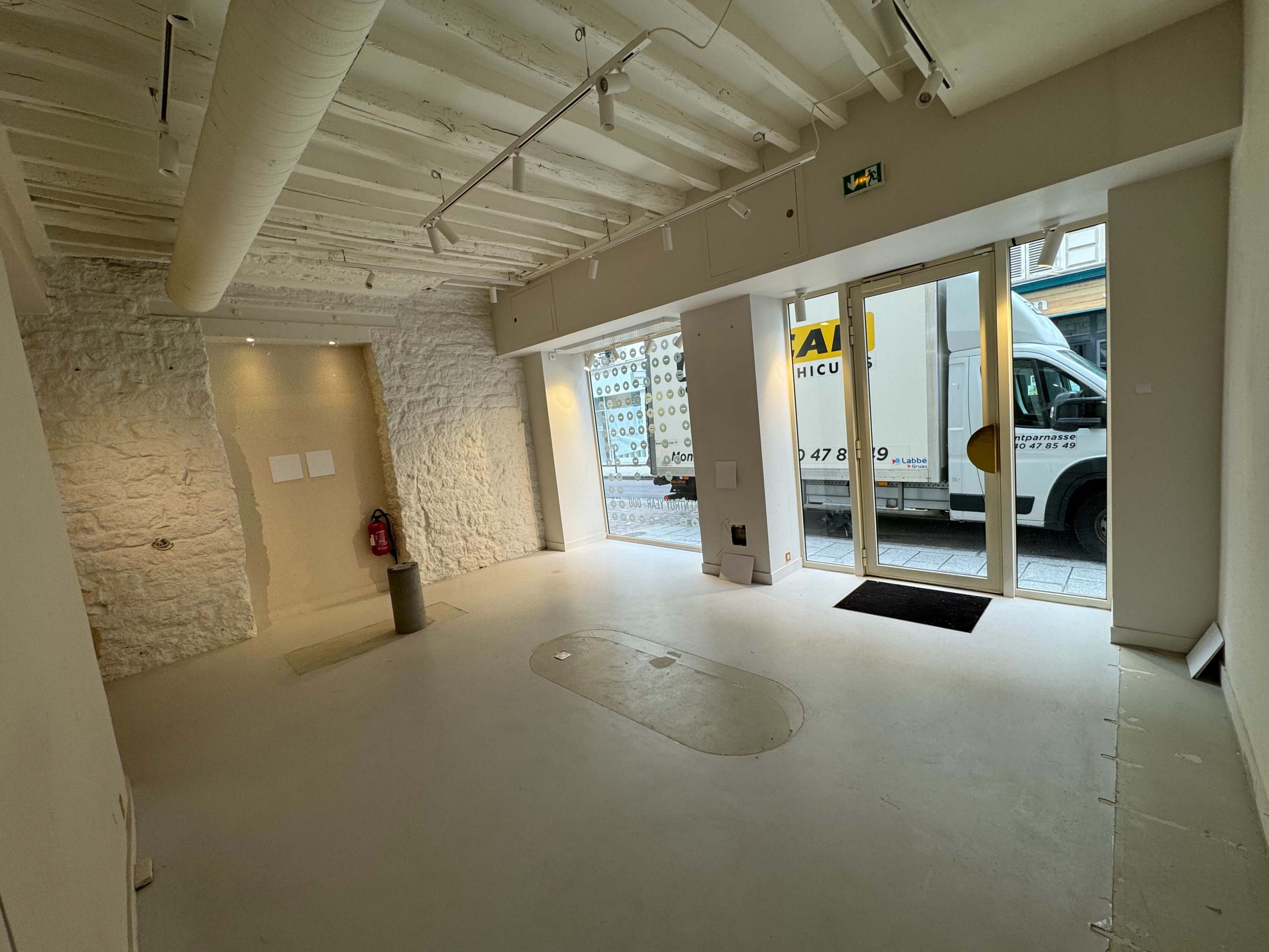 An empty commercial space with a stone wall, white ceiling beams, and large glass doors facing a street, while a moving truck is parked outside.