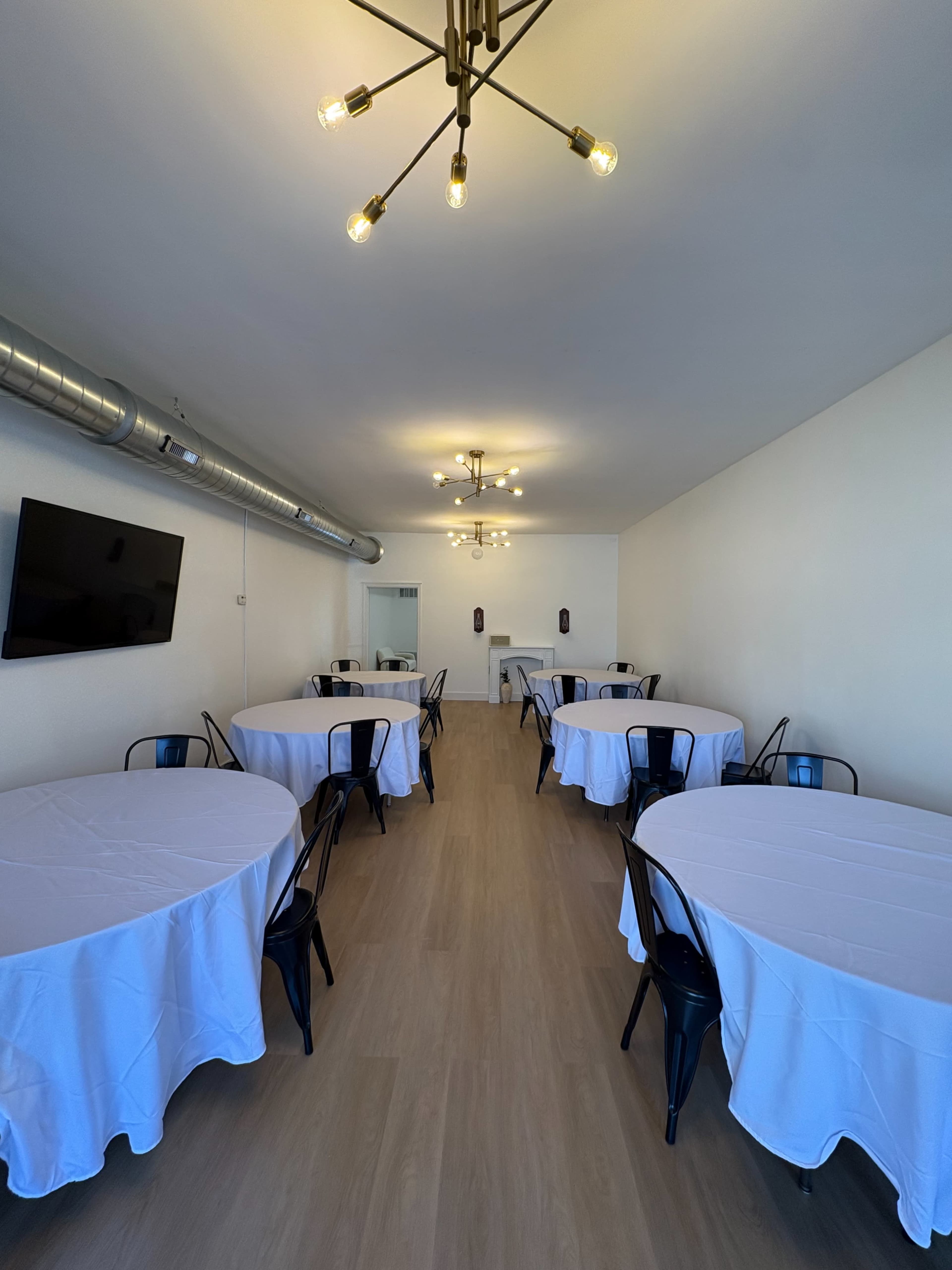 The image shows a spacious room with multiple round tables covered in white tablecloths, illuminated by pendant lights and equipped with a wall-mounted television.