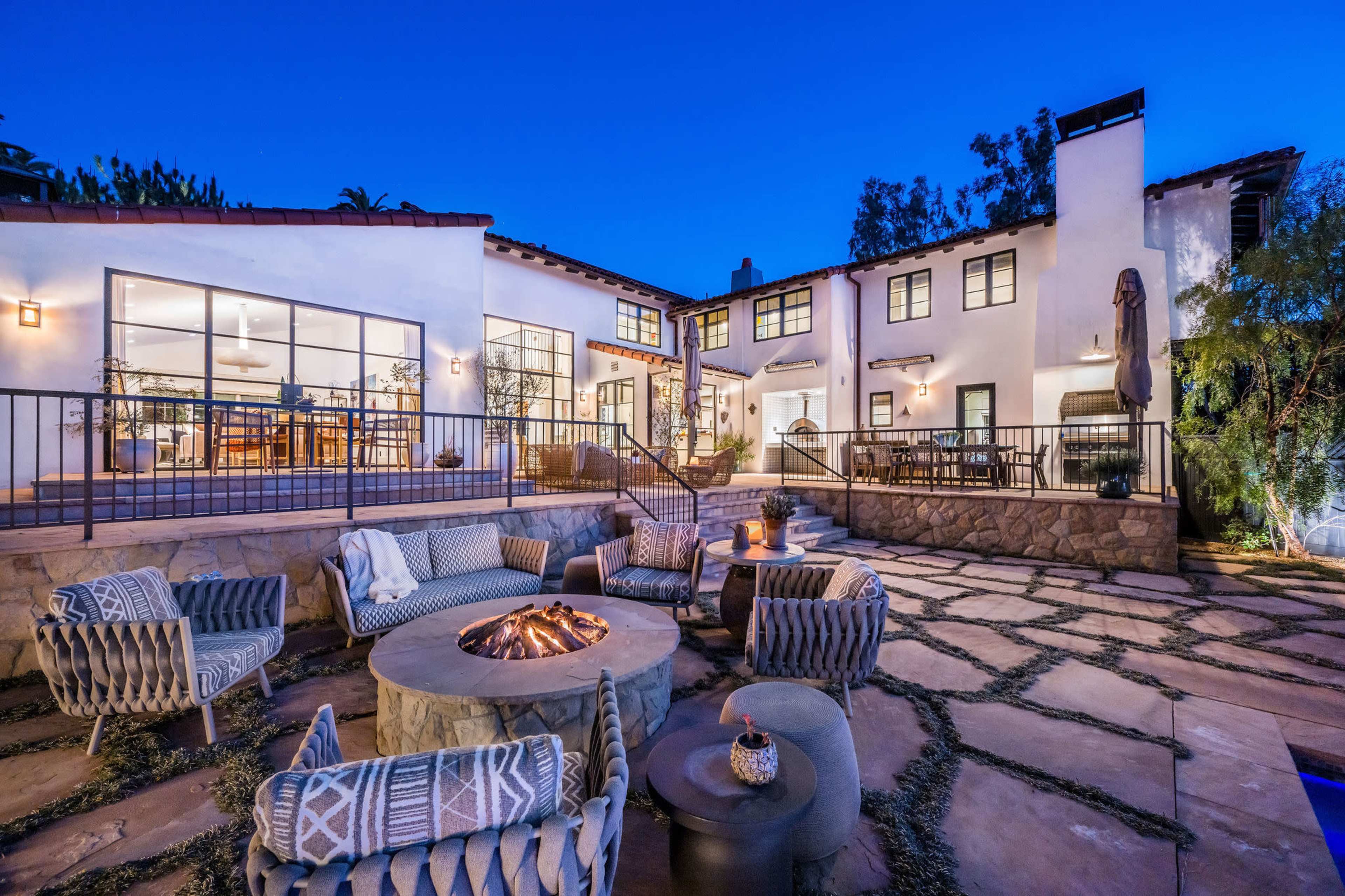A modern two-story house with large windows is surrounded by a stone patio featuring a fire pit and outdoor seating, set against a twilight sky.