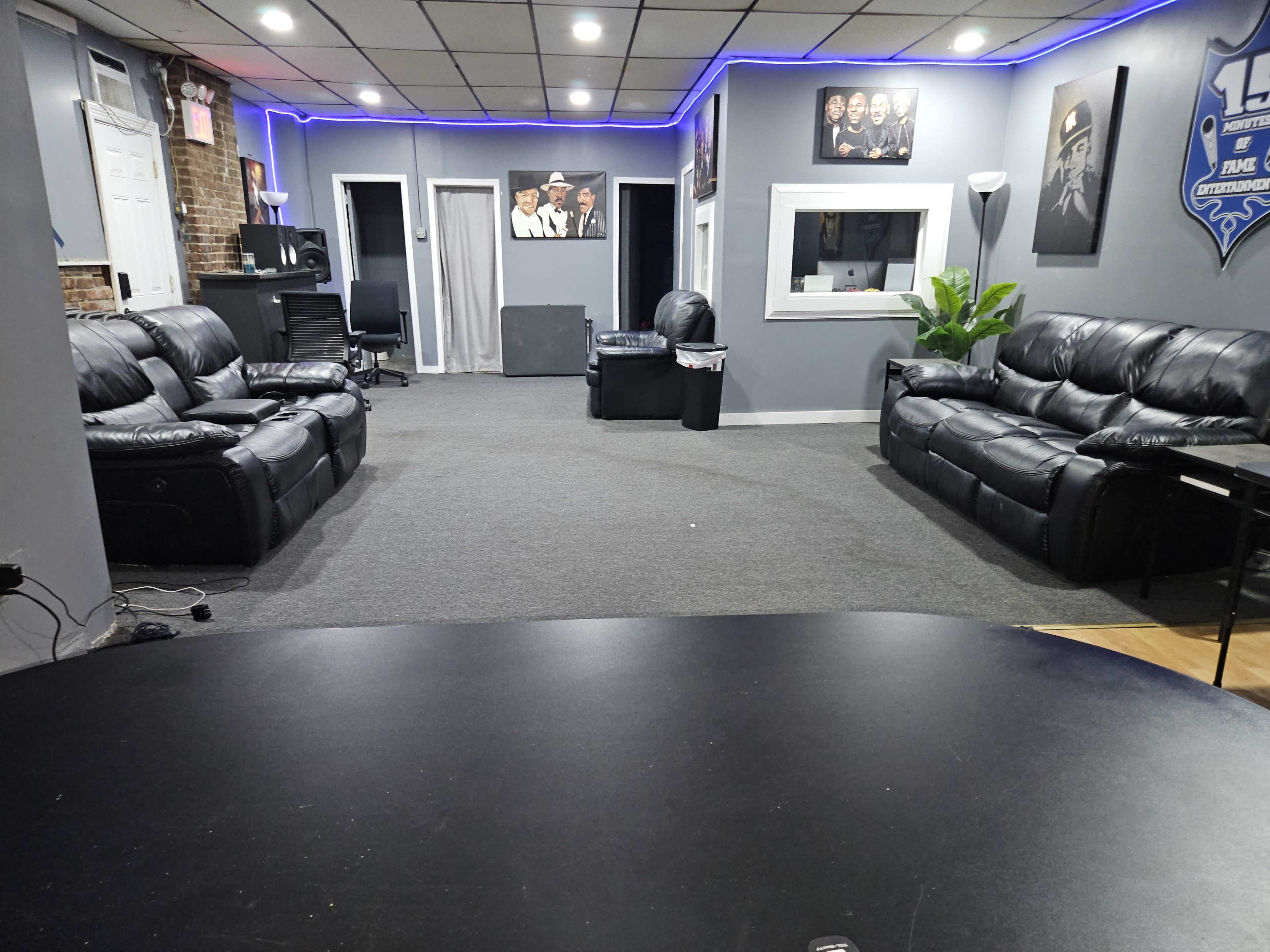 The image shows a spacious, well-lit room with black leather couches, a desk, and wall decorations, featuring a large window and contrasting flooring.