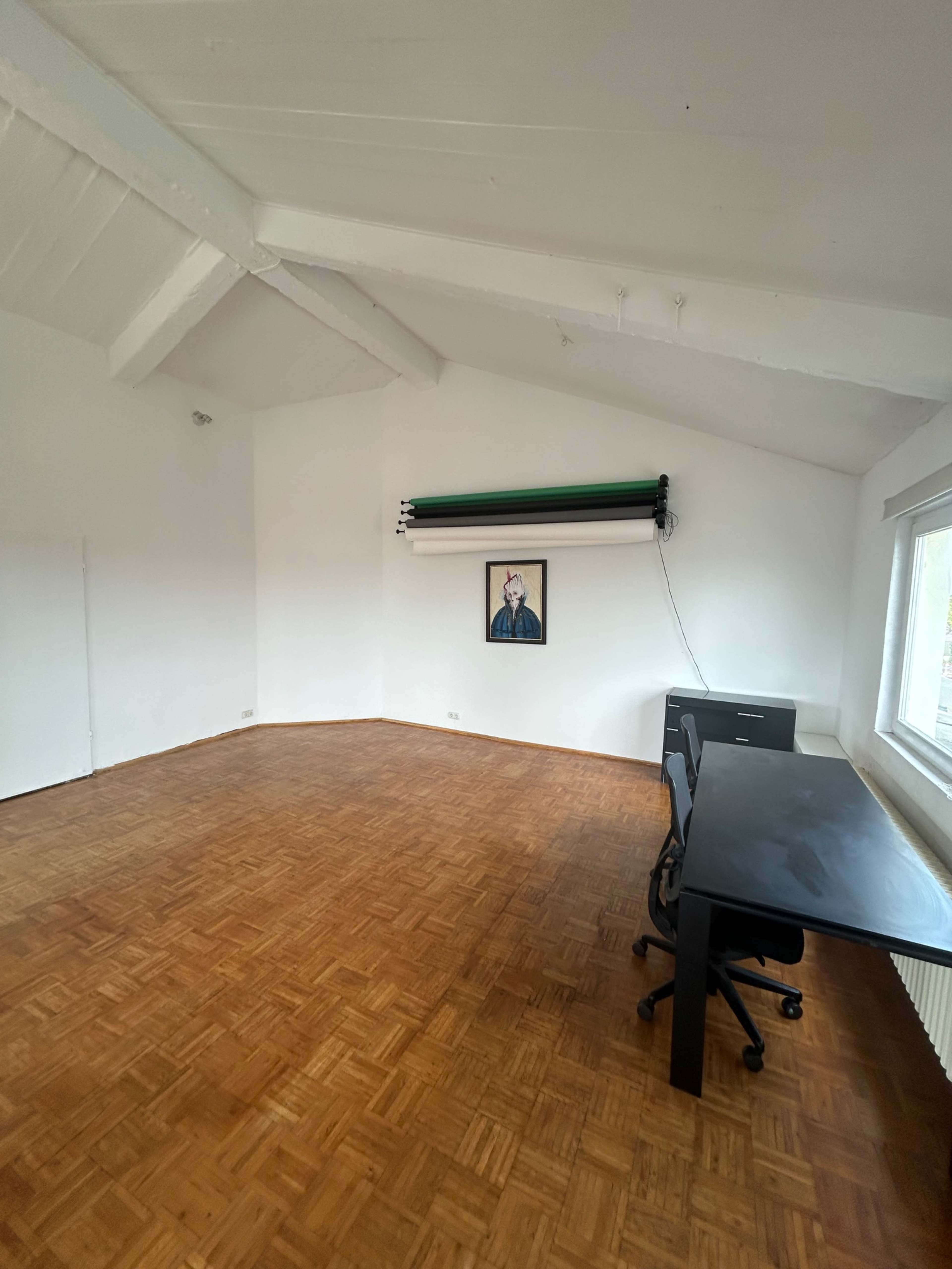 The image shows an empty room with a wooden floor, a large desk, a chair, and a framed portrait on the wall.