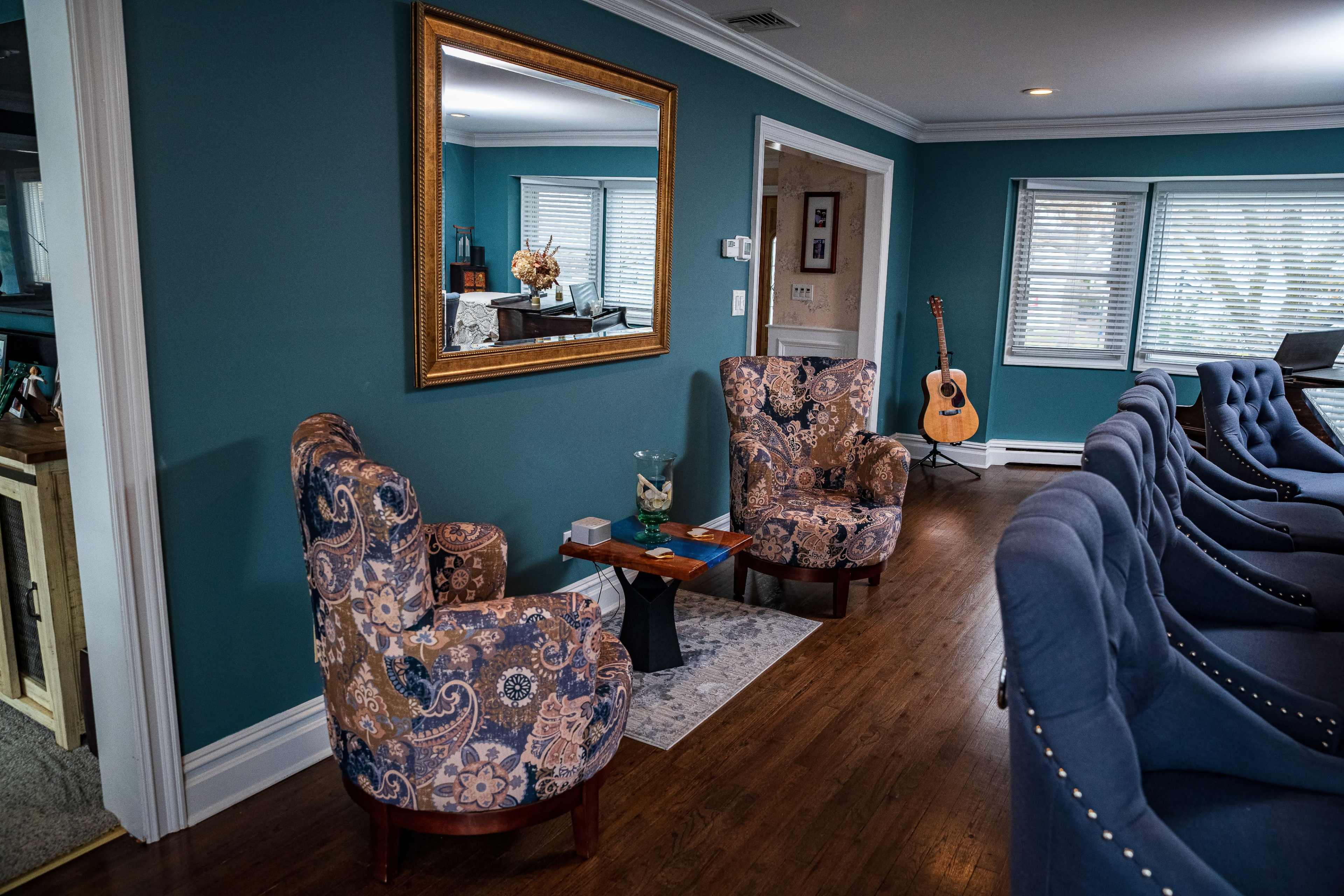 The image shows a cozy living room with two upholstered chairs, a small table between them, a large mirror on the wall, and a guitar in the corner.