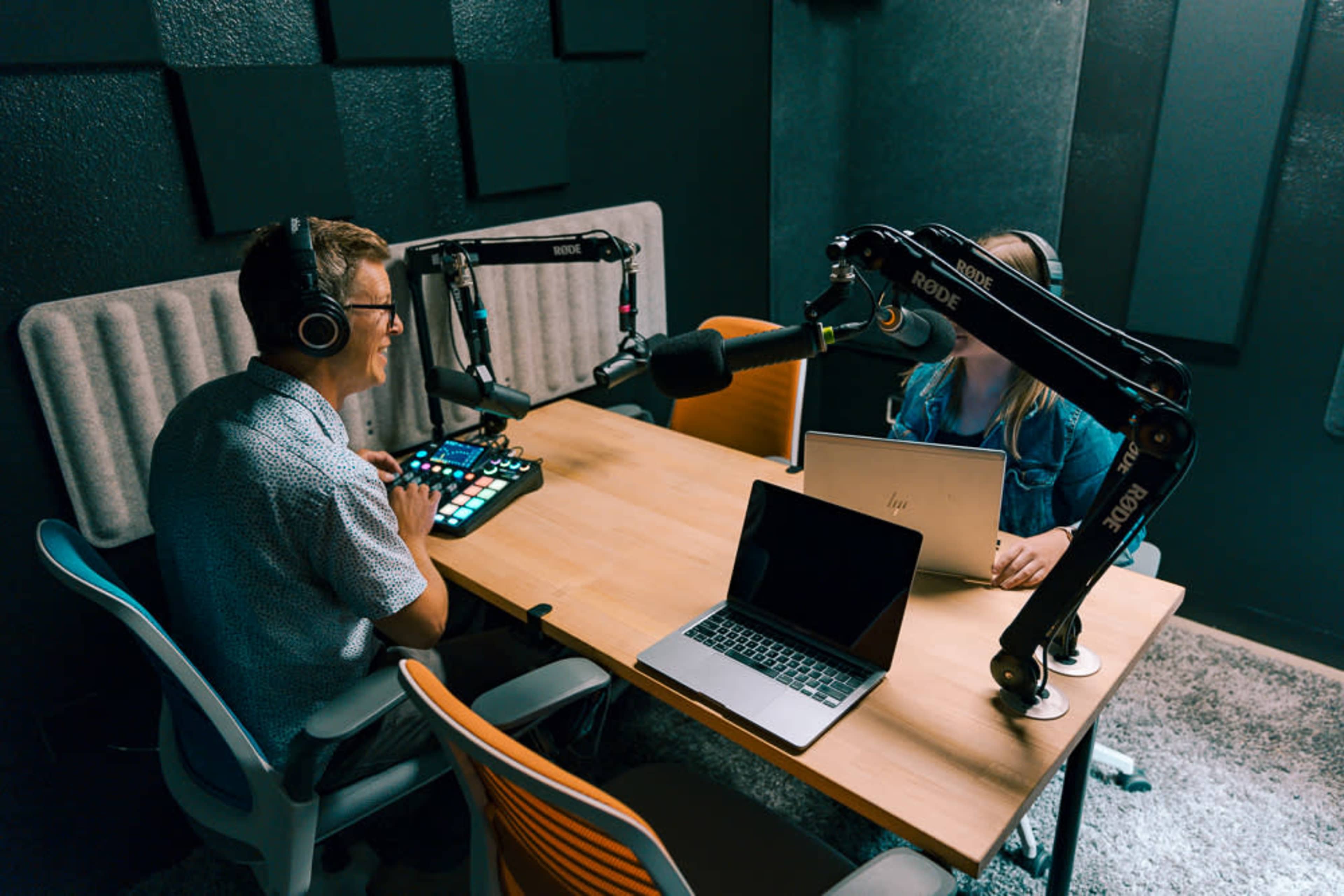 Two people sit at a table in a recording studio, equipped with microphones and laptops, preparing for a podcast session.