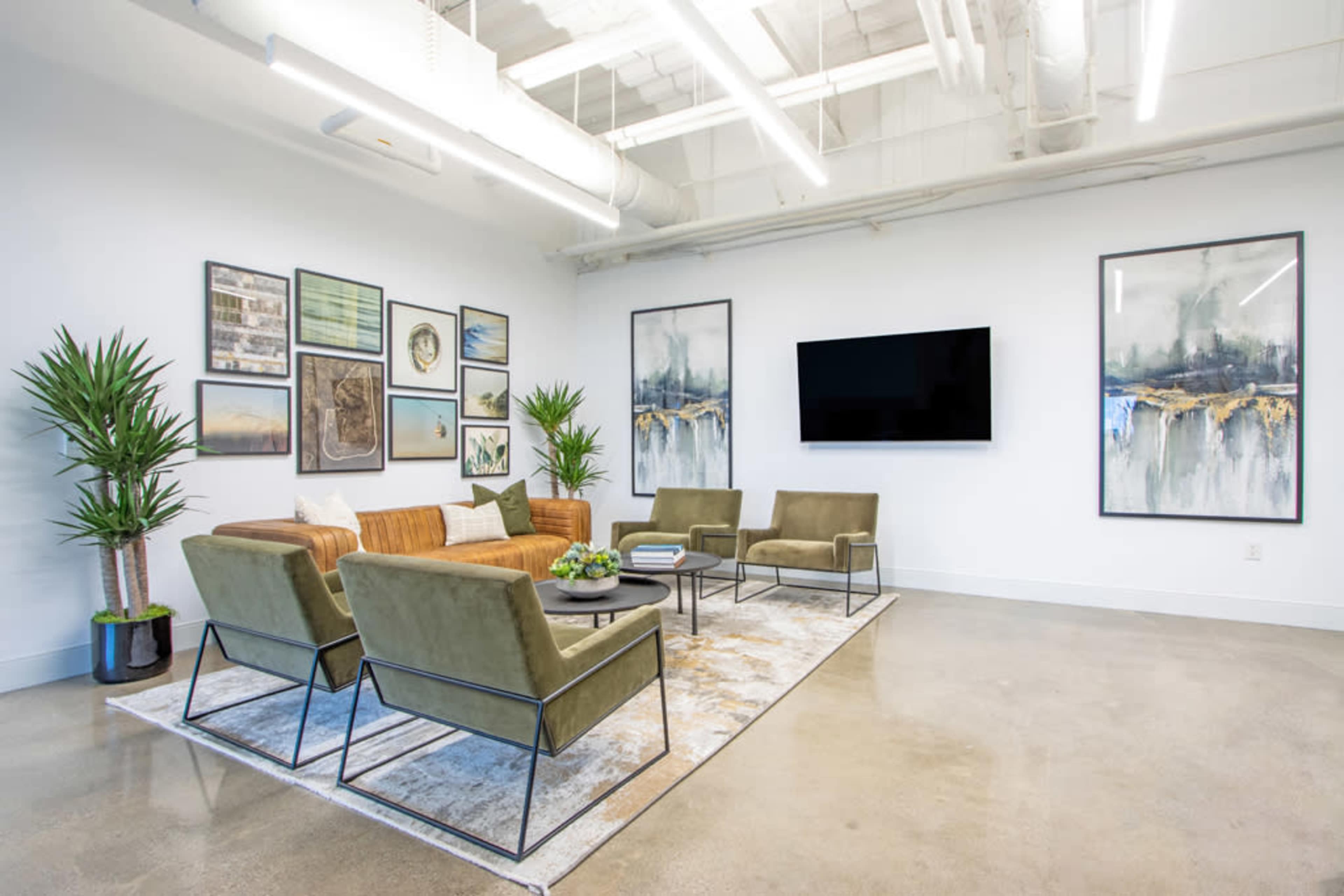 A modern lounge area features a brown sofa, green armchairs, a decorative rug, and framed artworks on the walls, with a television mounted centrally.