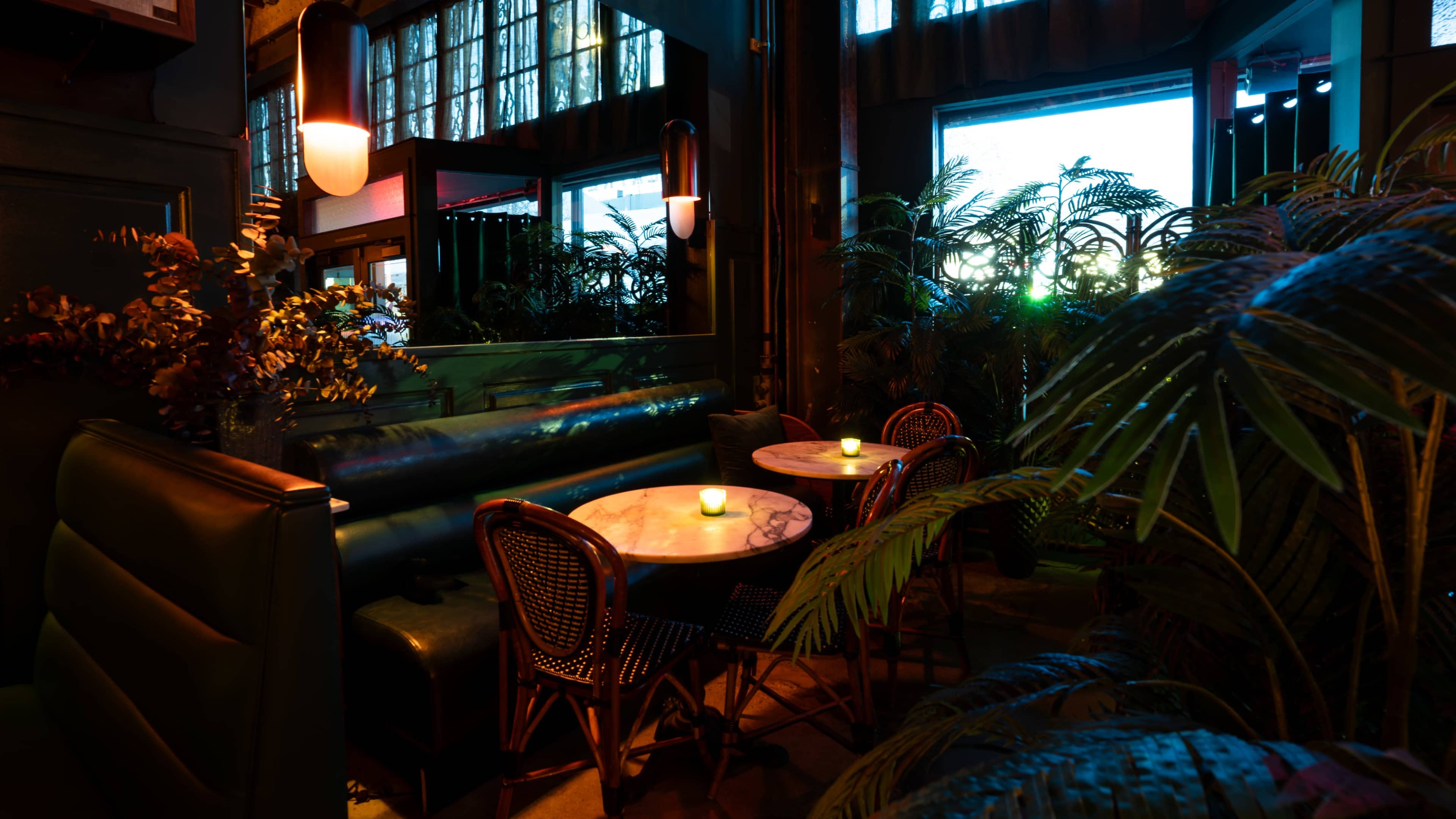 The image shows a cozy dining area with two tables surrounded by lush plants and soft lighting.
