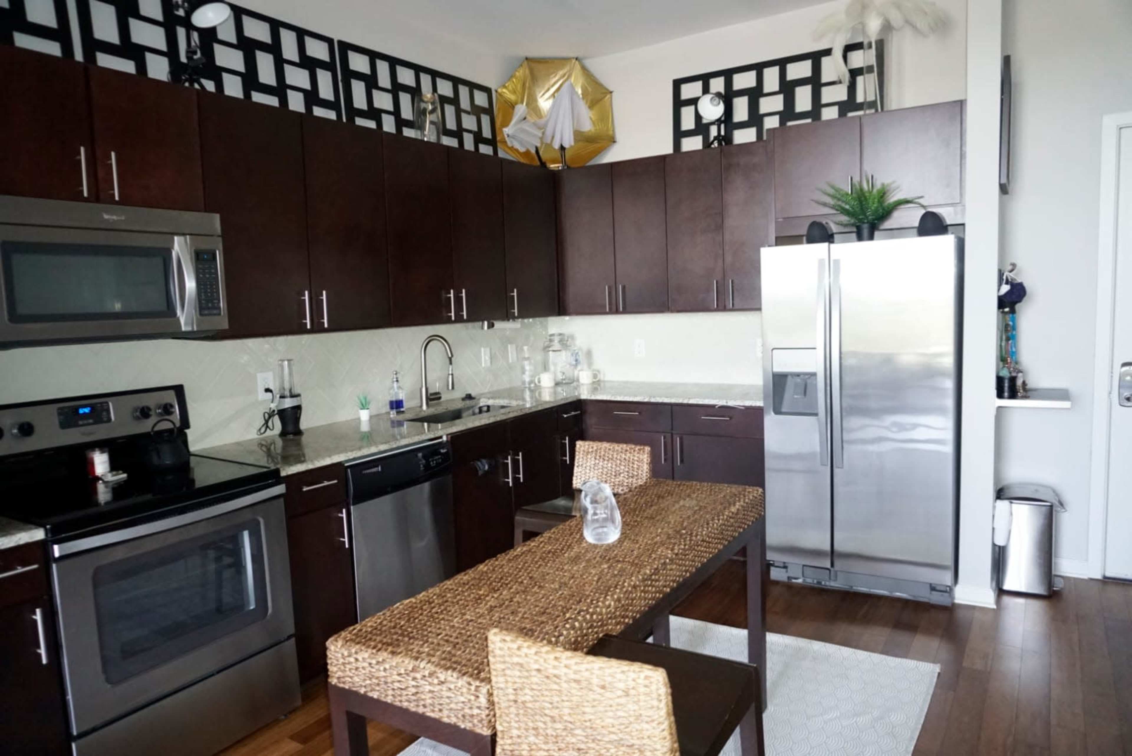 The image shows a modern kitchen with dark wooden cabinets, stainless steel appliances, and a small dining table set with wicker chairs.