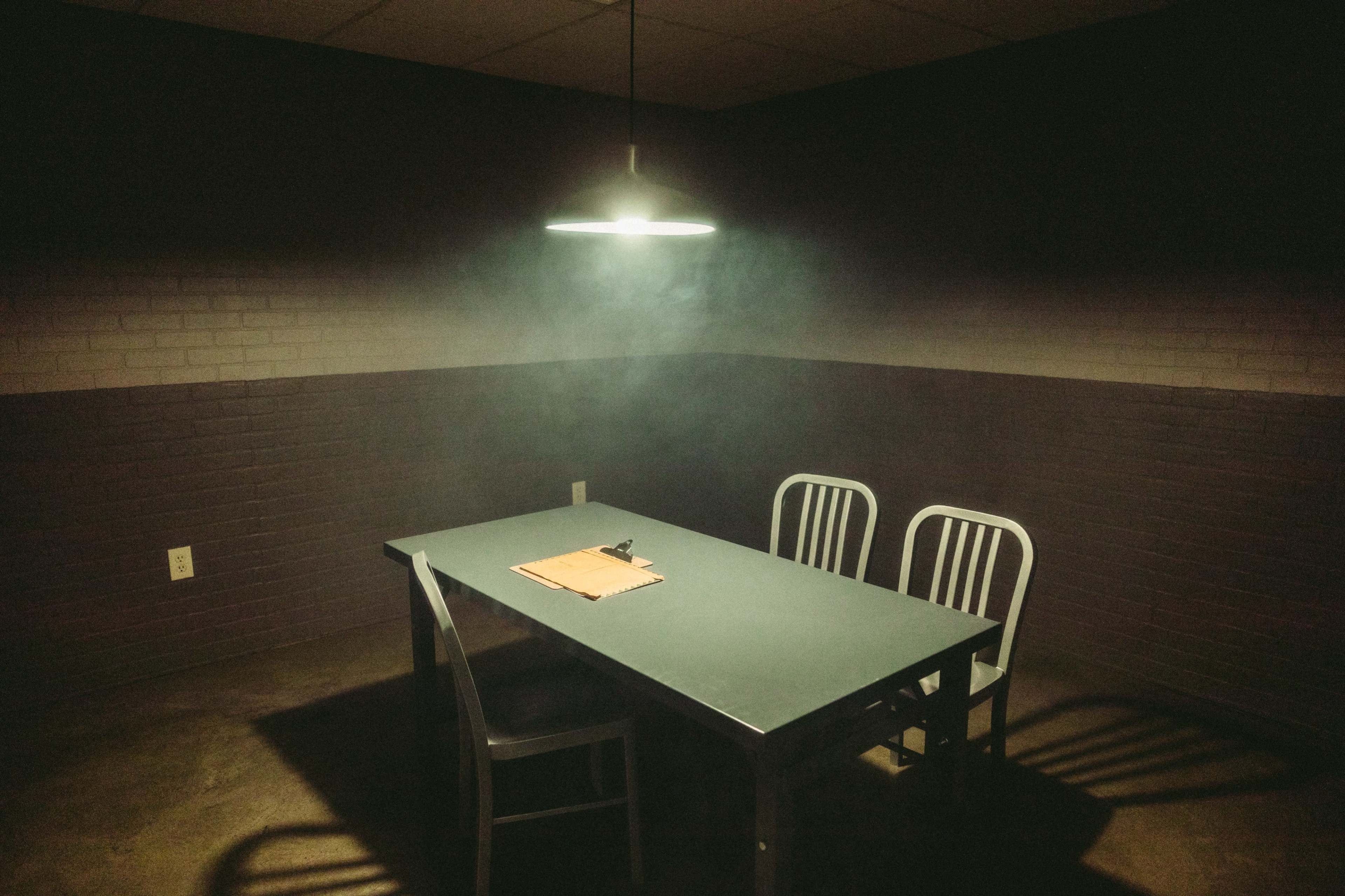 A dimly lit interrogation room features a gray table, three metal chairs, and a clipboard resting on the table.