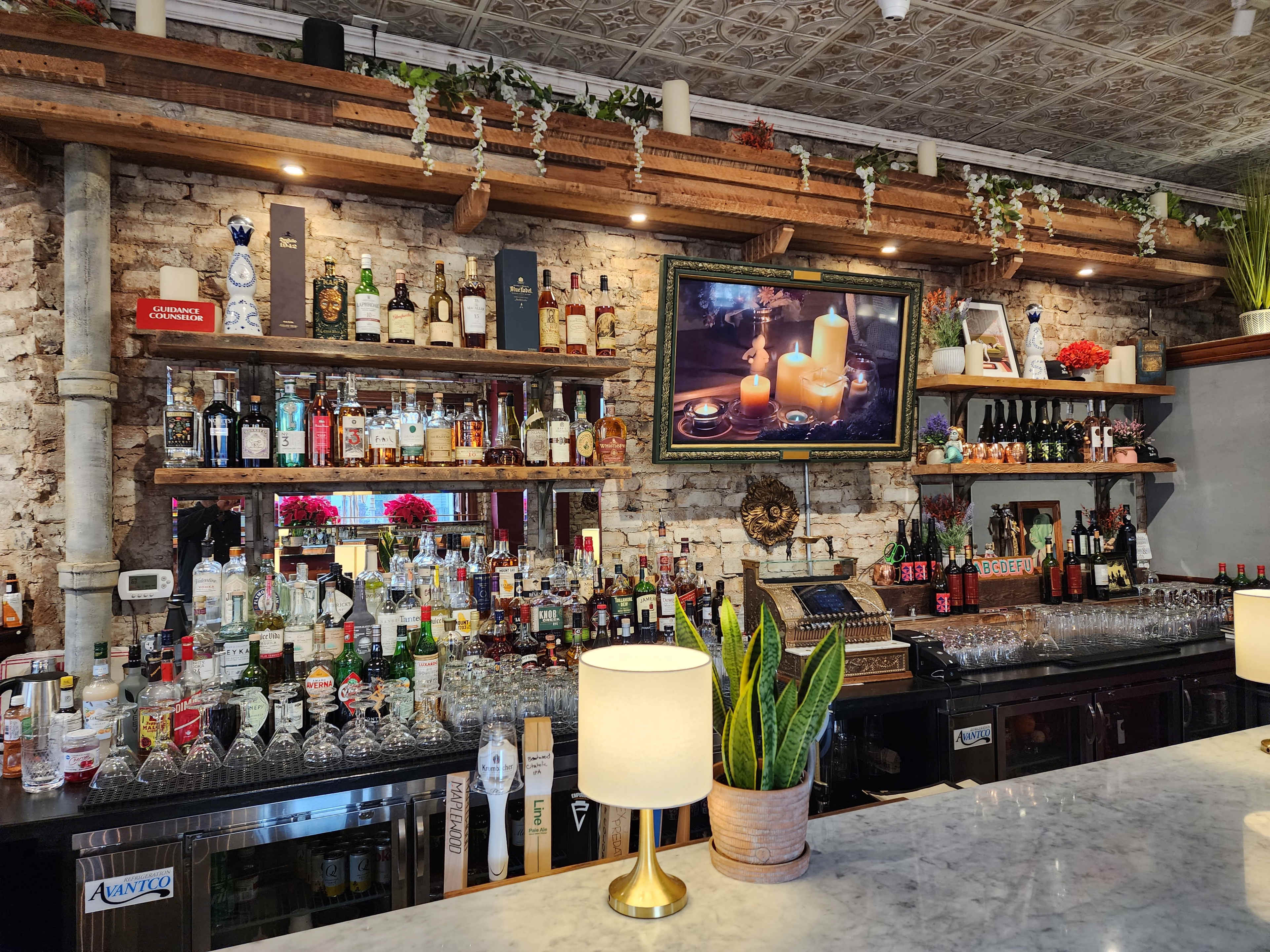 The image shows a well-stocked bar with a wooden countertop, shelves filled with various liquor bottles, and a television displaying a candlelit scene above the bar.