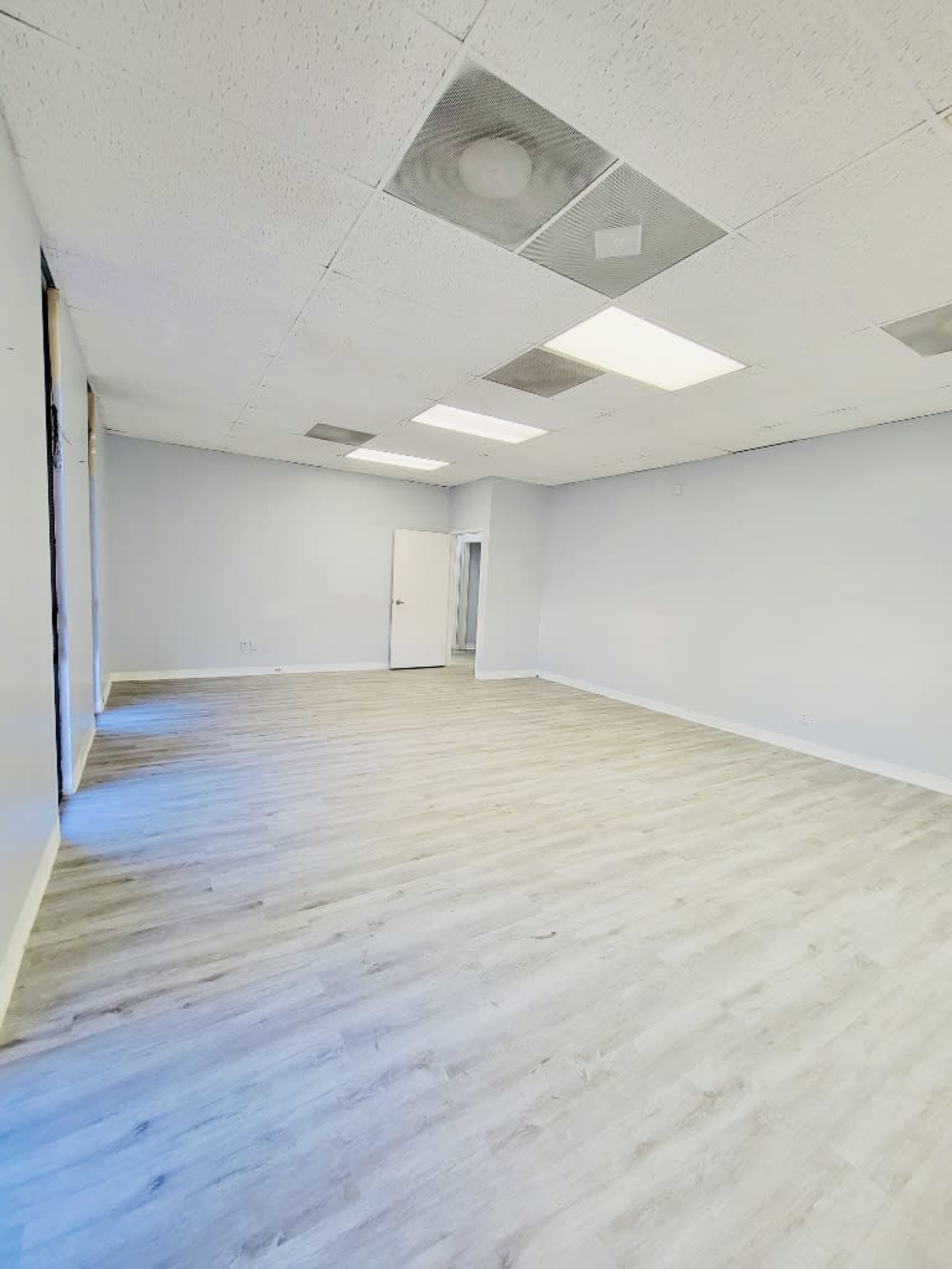 A vacant room with light-colored walls, a light wooden floor, and several ceiling lights.