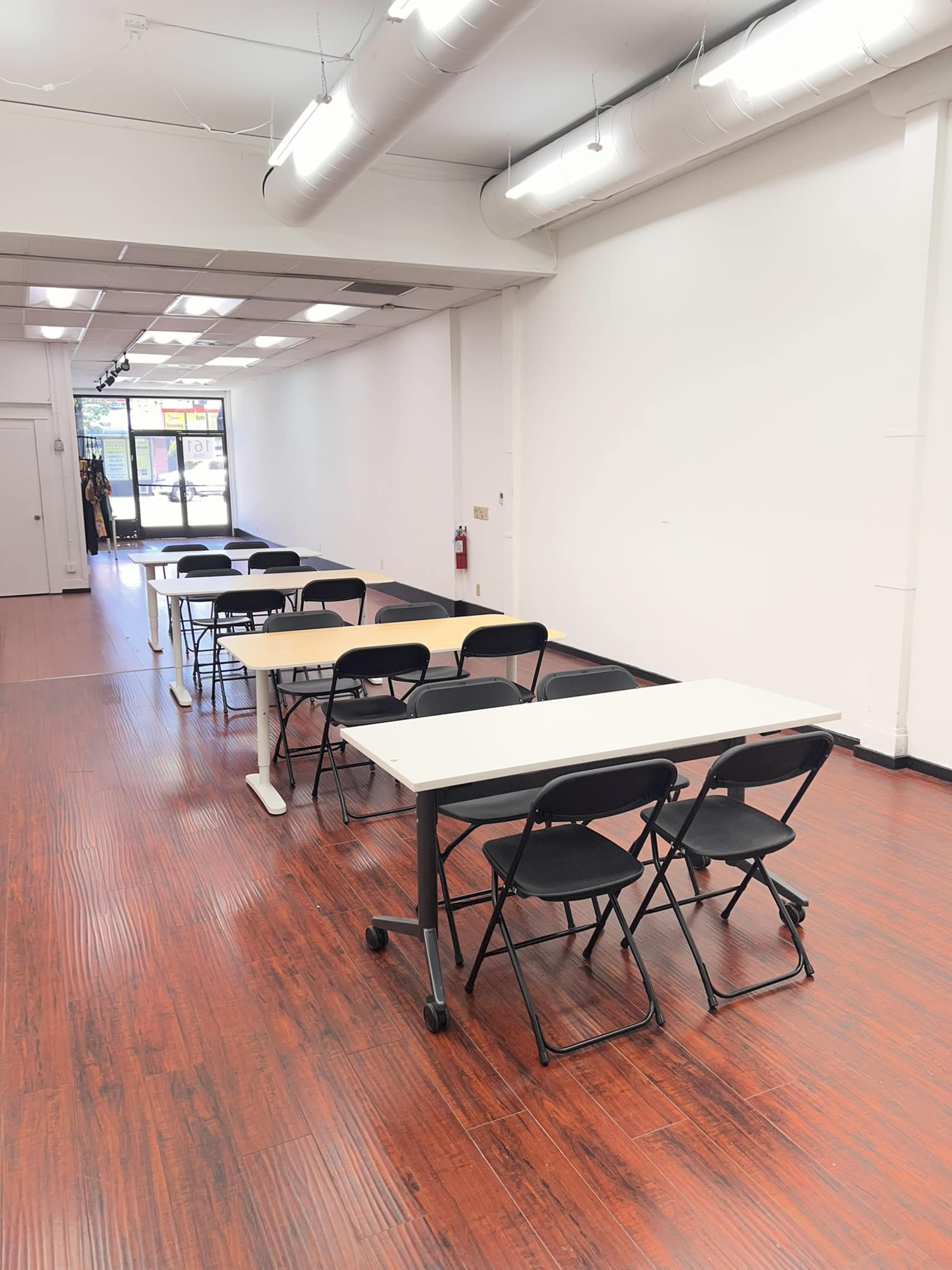 The image shows an empty room with several rows of tables and chairs set up along the wooden floor, illuminated by overhead lights.