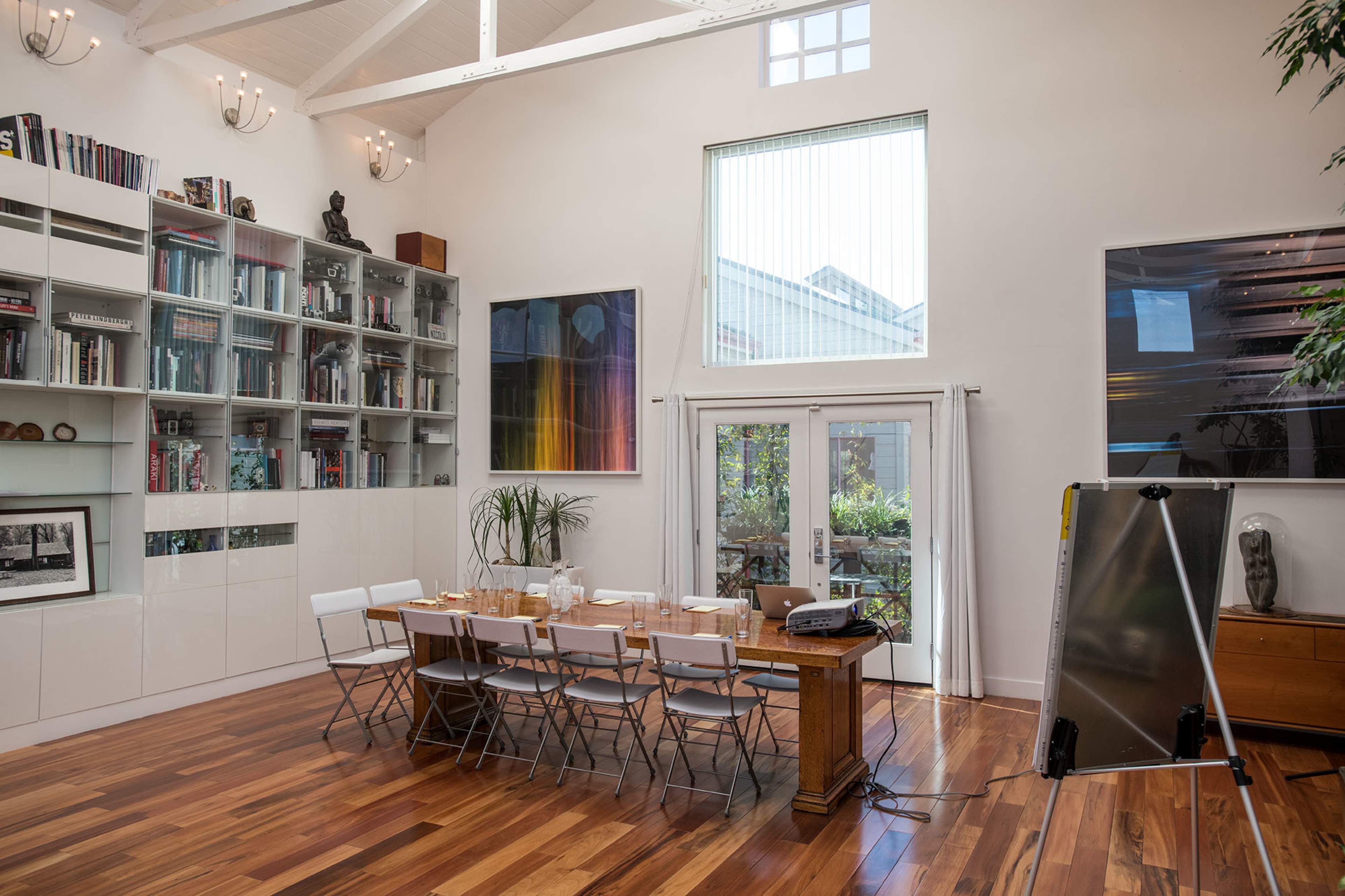 The image shows a bright, spacious room with wooden flooring, a long dining table set for a meal, bookshelves, and large windows allowing natural light to enter.