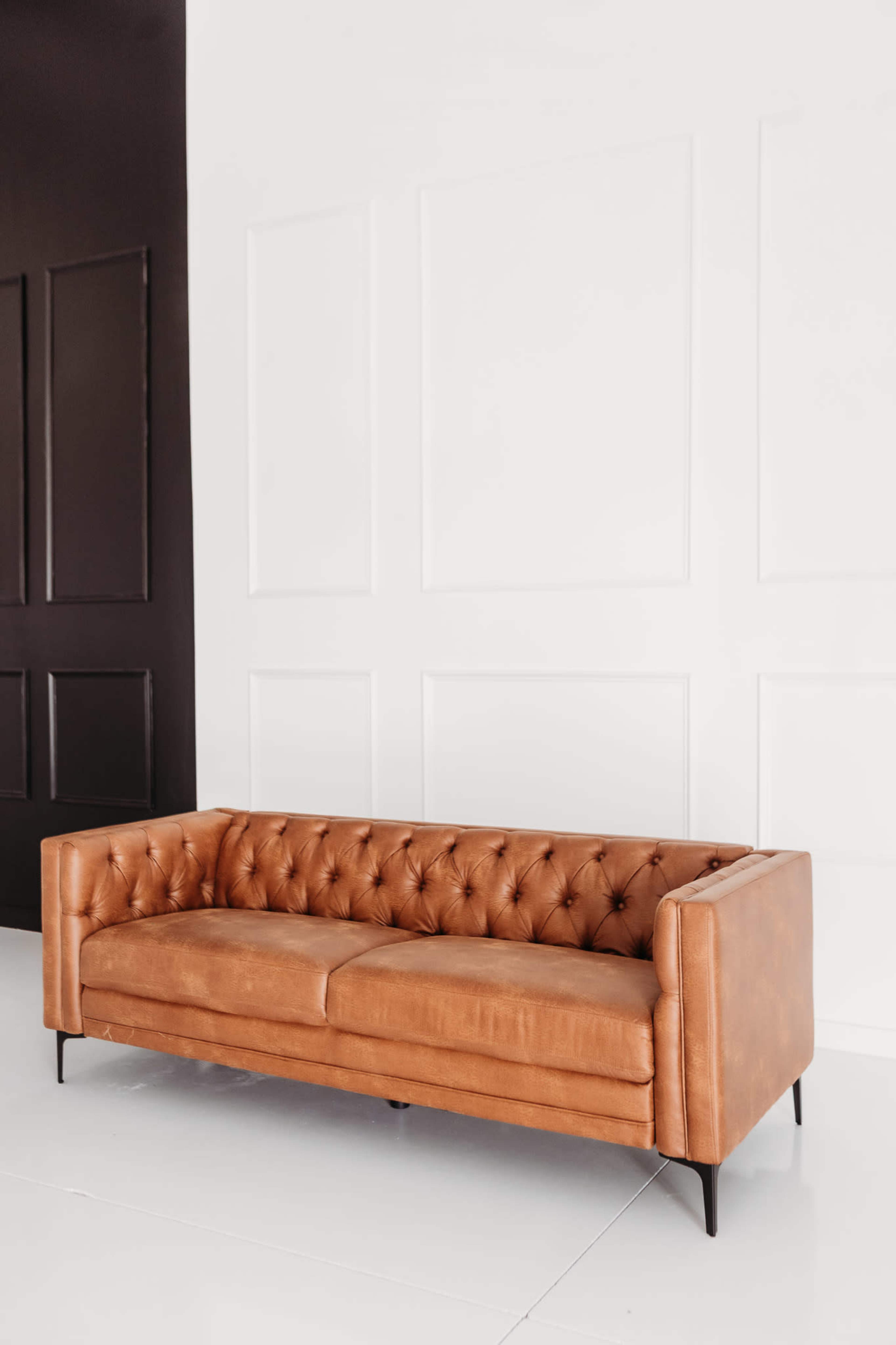 A tan leather couch with button tufting is placed against a white wall, juxtaposed with a dark panel.