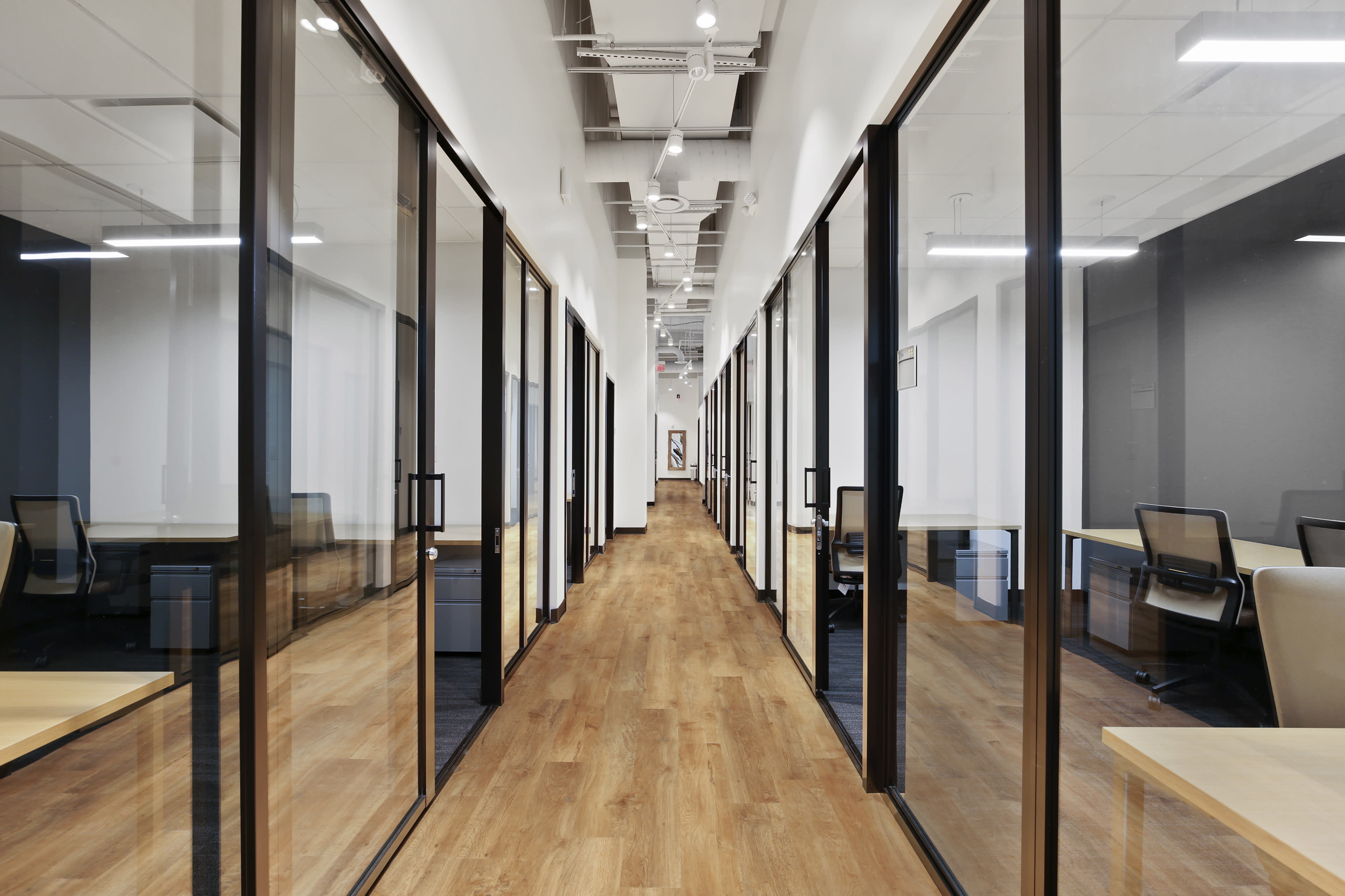 The image shows a modern office hallway lined with glass-walled meeting rooms on either side, featuring wooden flooring and bright overhead lights.