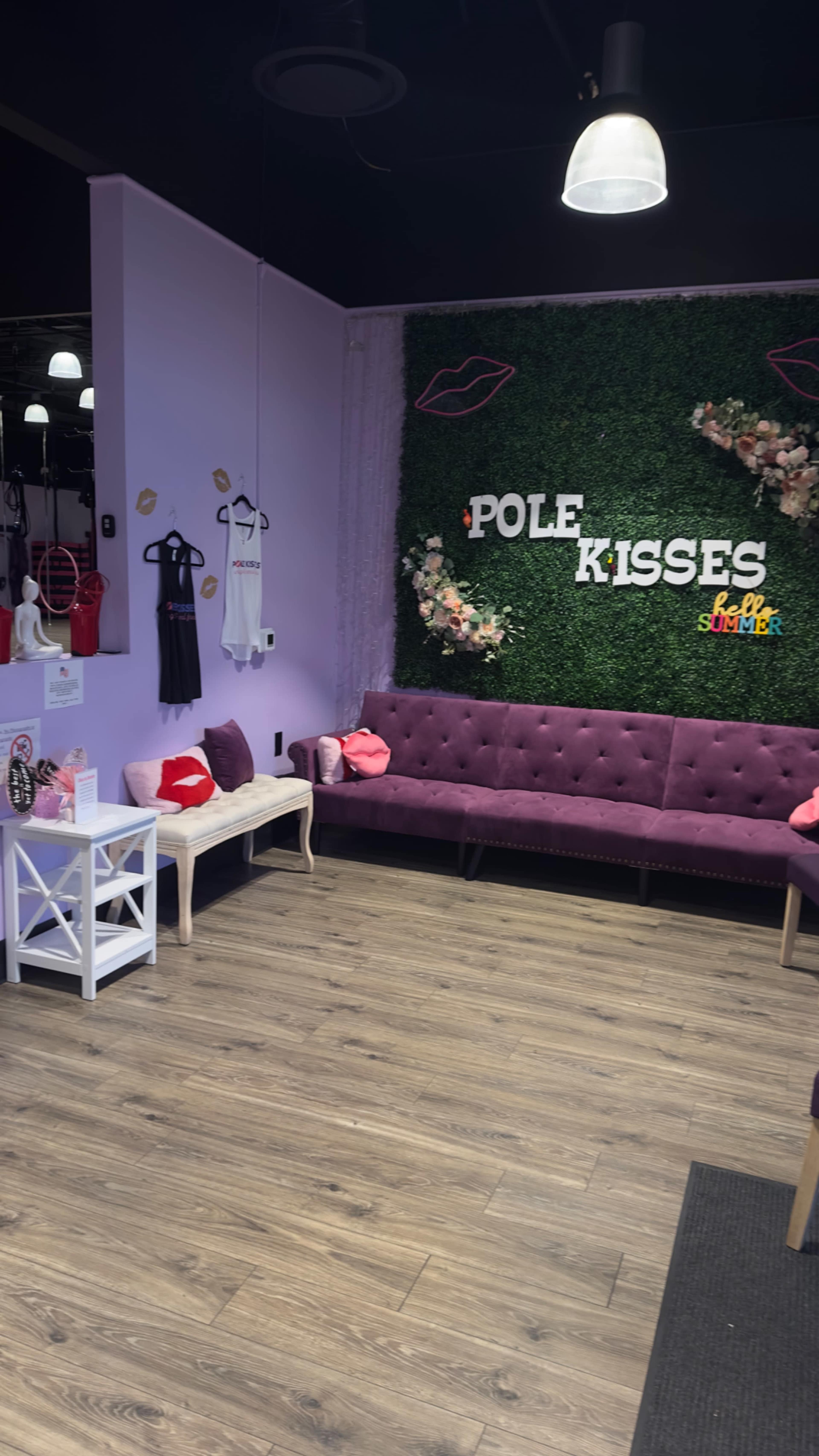 A brightly colored shop interior with a purple sofa, decorative wall plants spelling "Pole Kisses," and various clothing items on display.