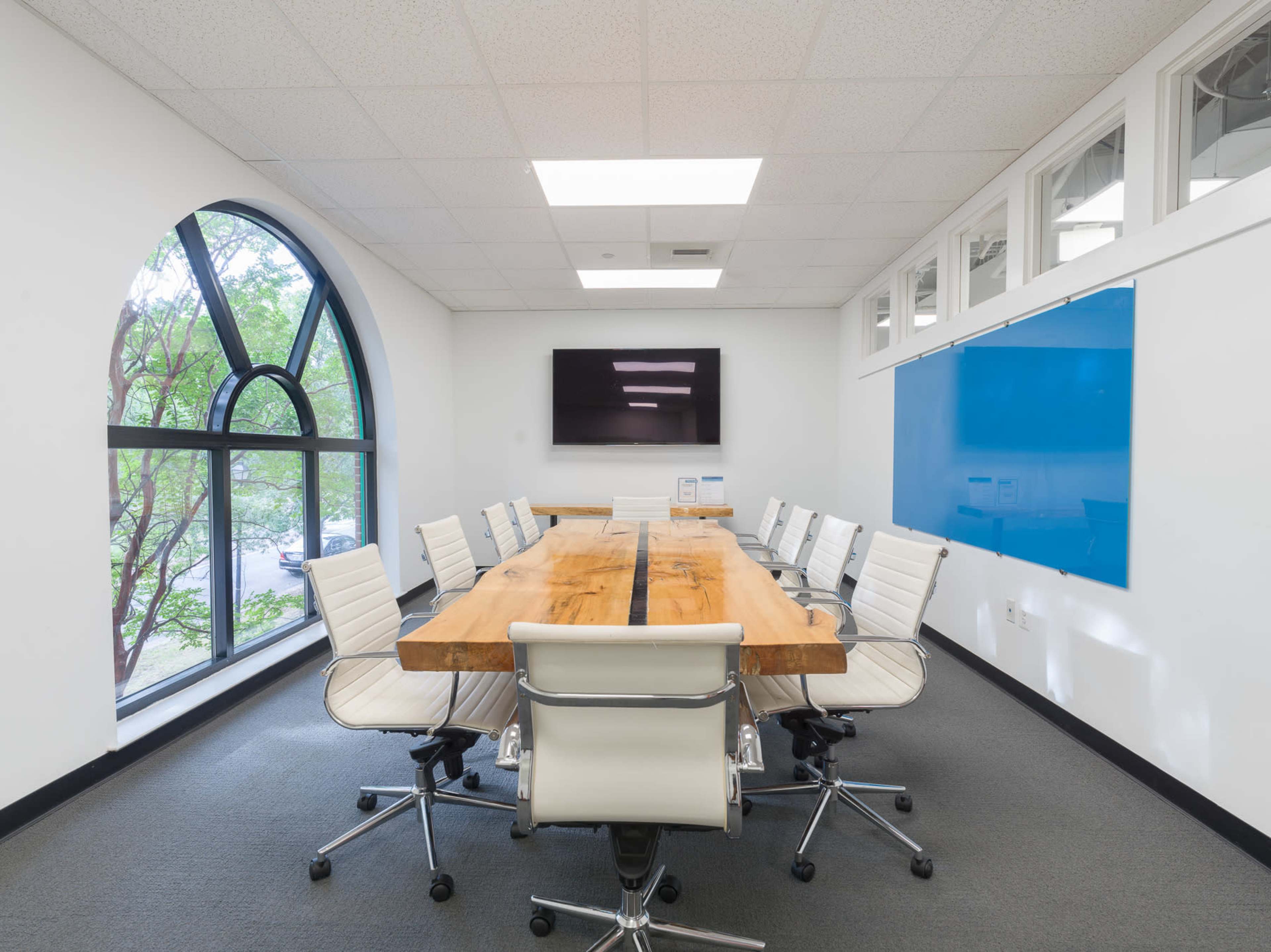 A conference room features a large wooden table surrounded by white leather chairs, with a large window and a wall-mounted screen.
