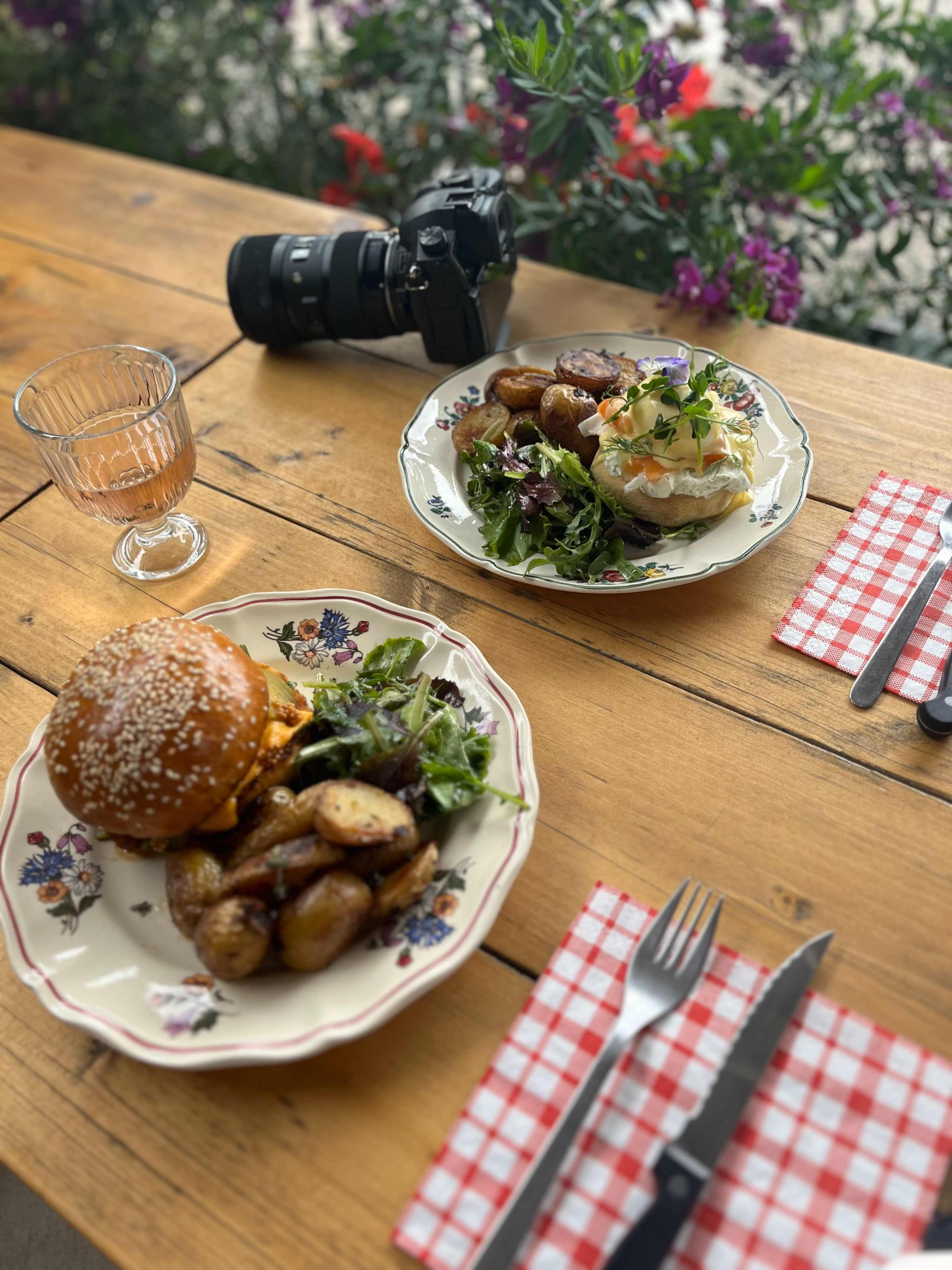 The image shows two plates of food on a wooden table, one featuring a sesame seed burger with salad and roasted potatoes, and the other a dish with salad, a soft egg, and garnishes, alongside a glass and a camera.