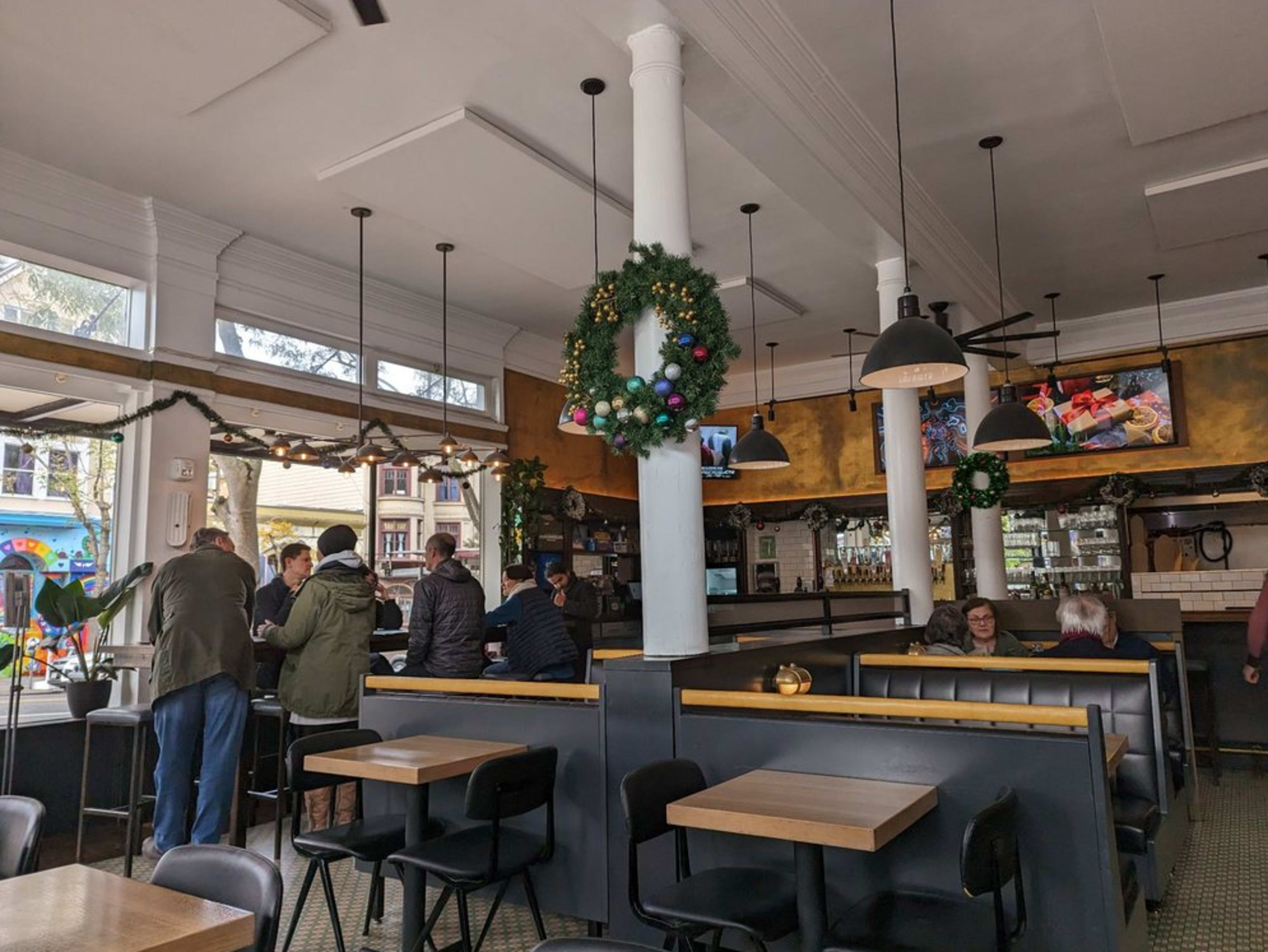 A bustling café interior features patrons conversing at tables, adorned with festive wreaths and colorful decorations.