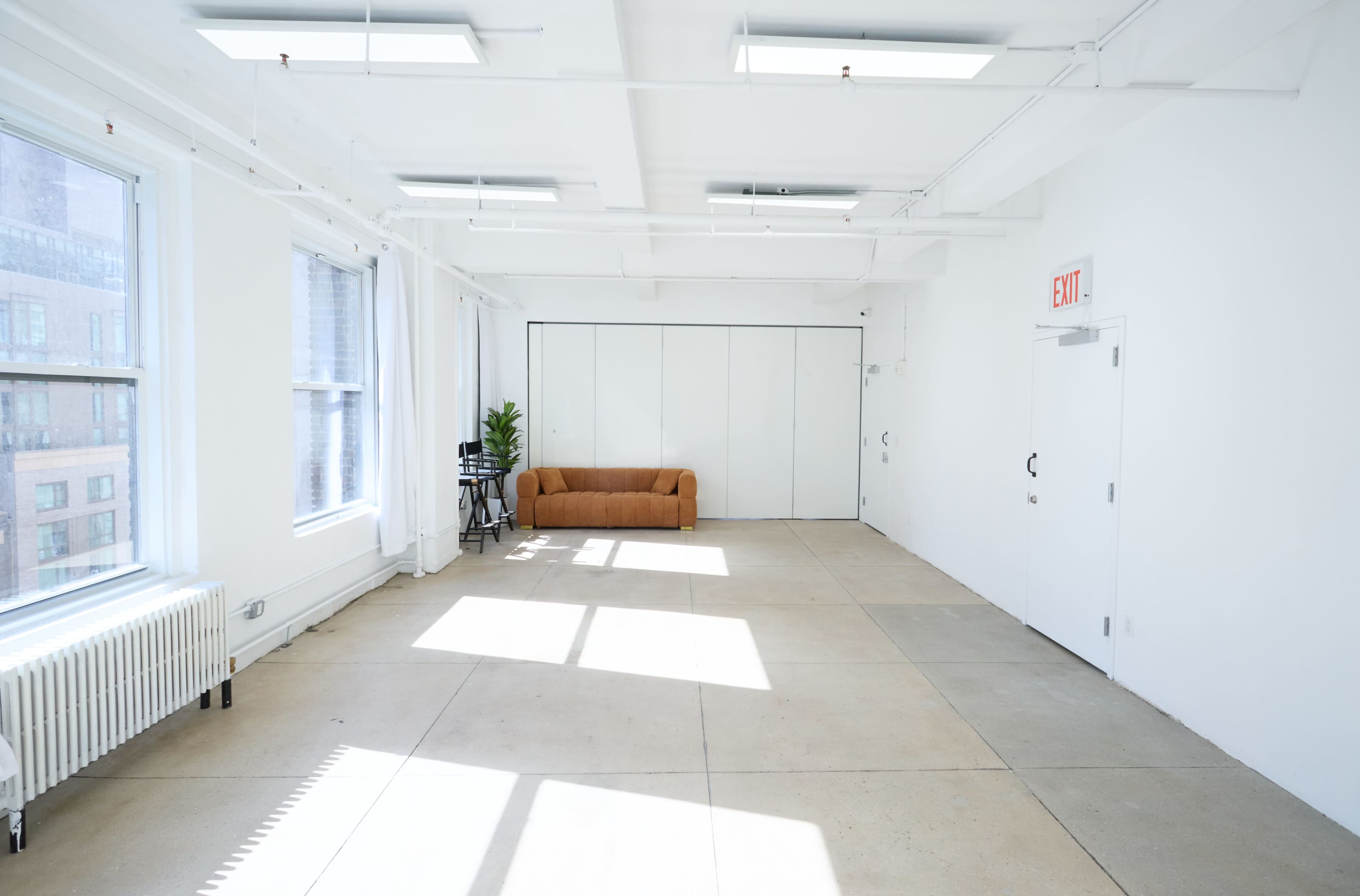 The image shows a bright, spacious room with large windows, a brown couch against one wall, and minimal furnishings.