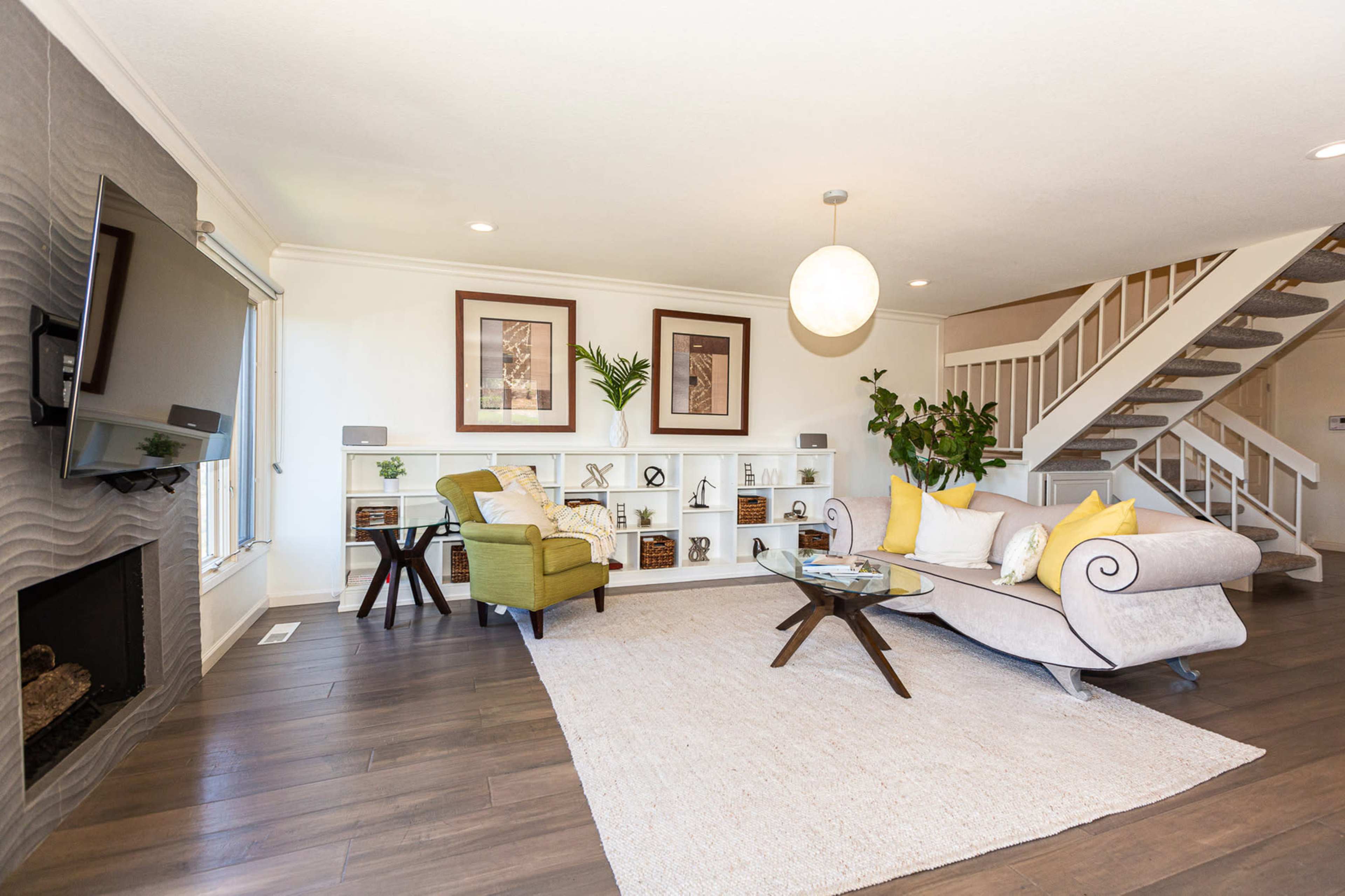 The living room features a light-colored sofa, an armchair, a coffee table, and a staircase, all accented by a large area rug and decorative shelves.