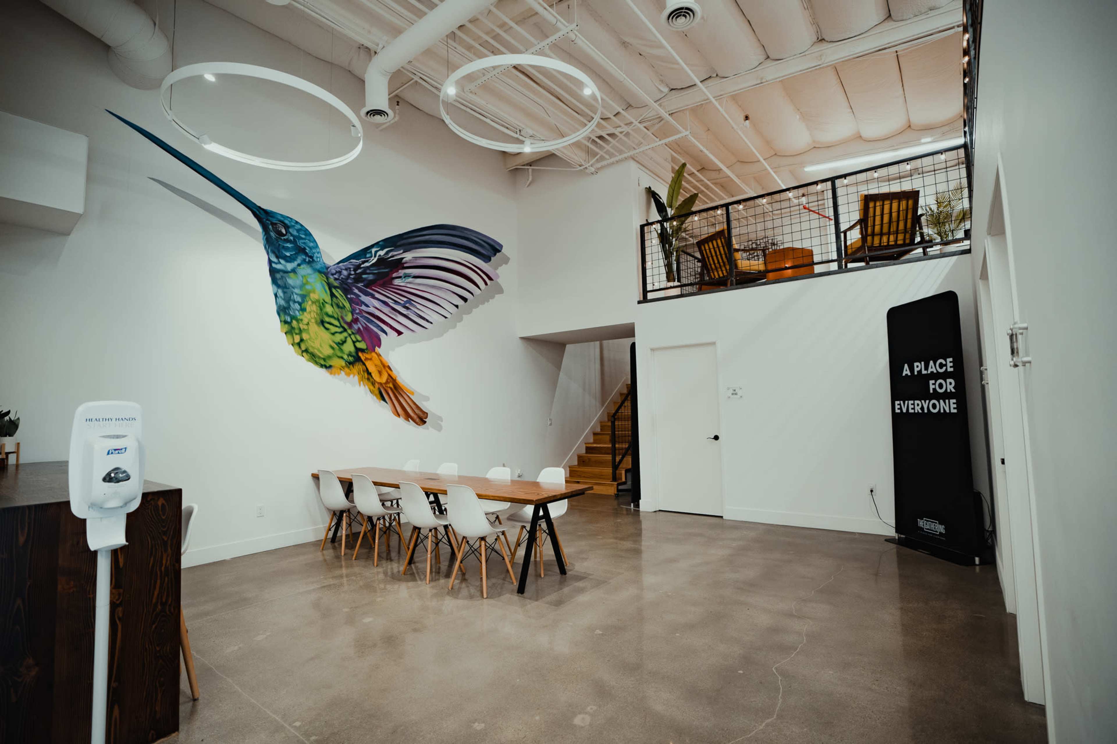 A spacious room with a large mural of a hummingbird on the wall, a long dining table with chairs in the center, and a staircase leading to an upper level.