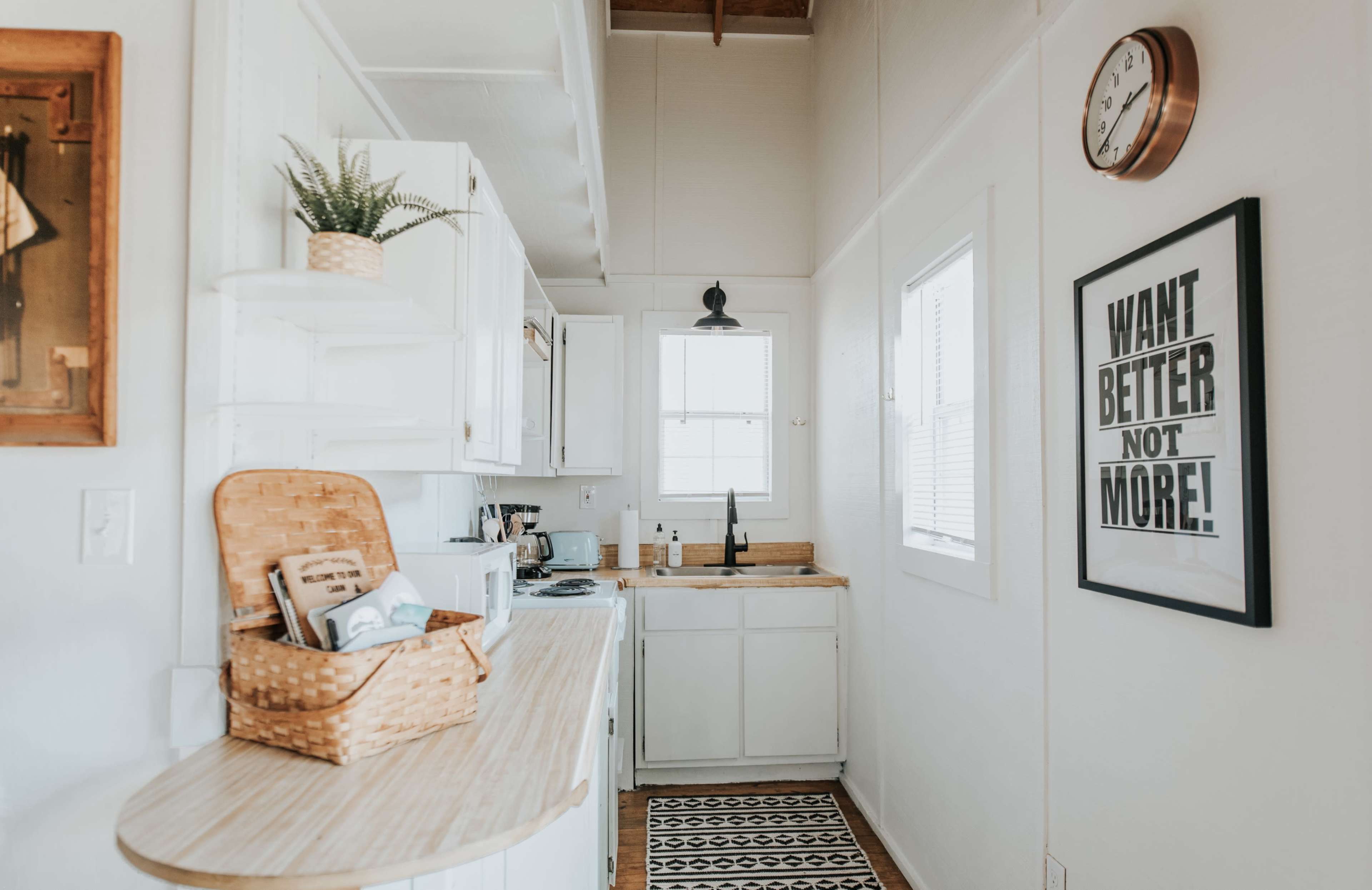 A cozy kitchen features white cabinets, a sink under a window, and minimal decor, including a wall clock and a framed motivational poster.