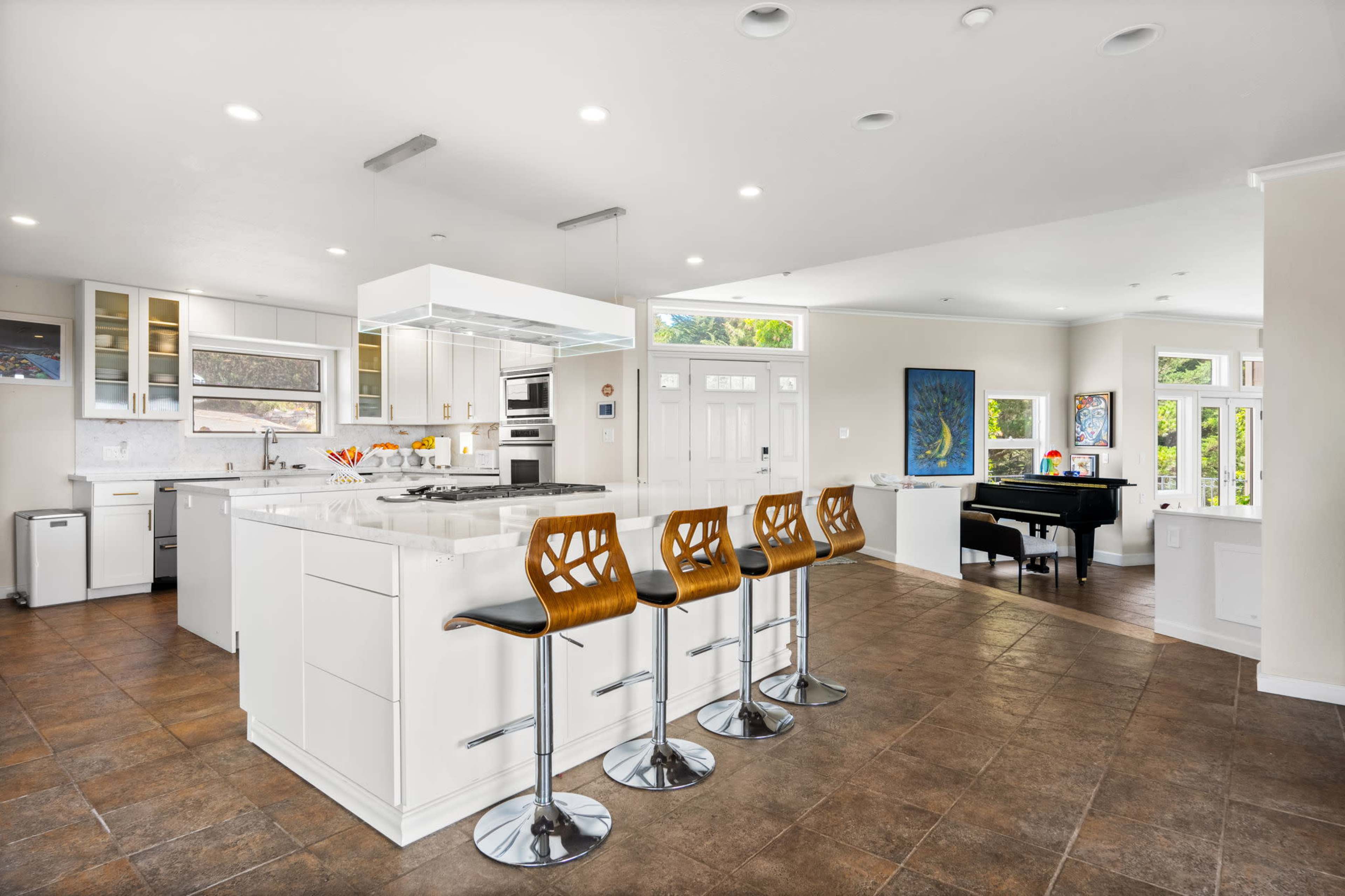 A modern kitchen with white cabinetry, a large island with bar stools, and an adjoining living area featuring a piano and large windows.