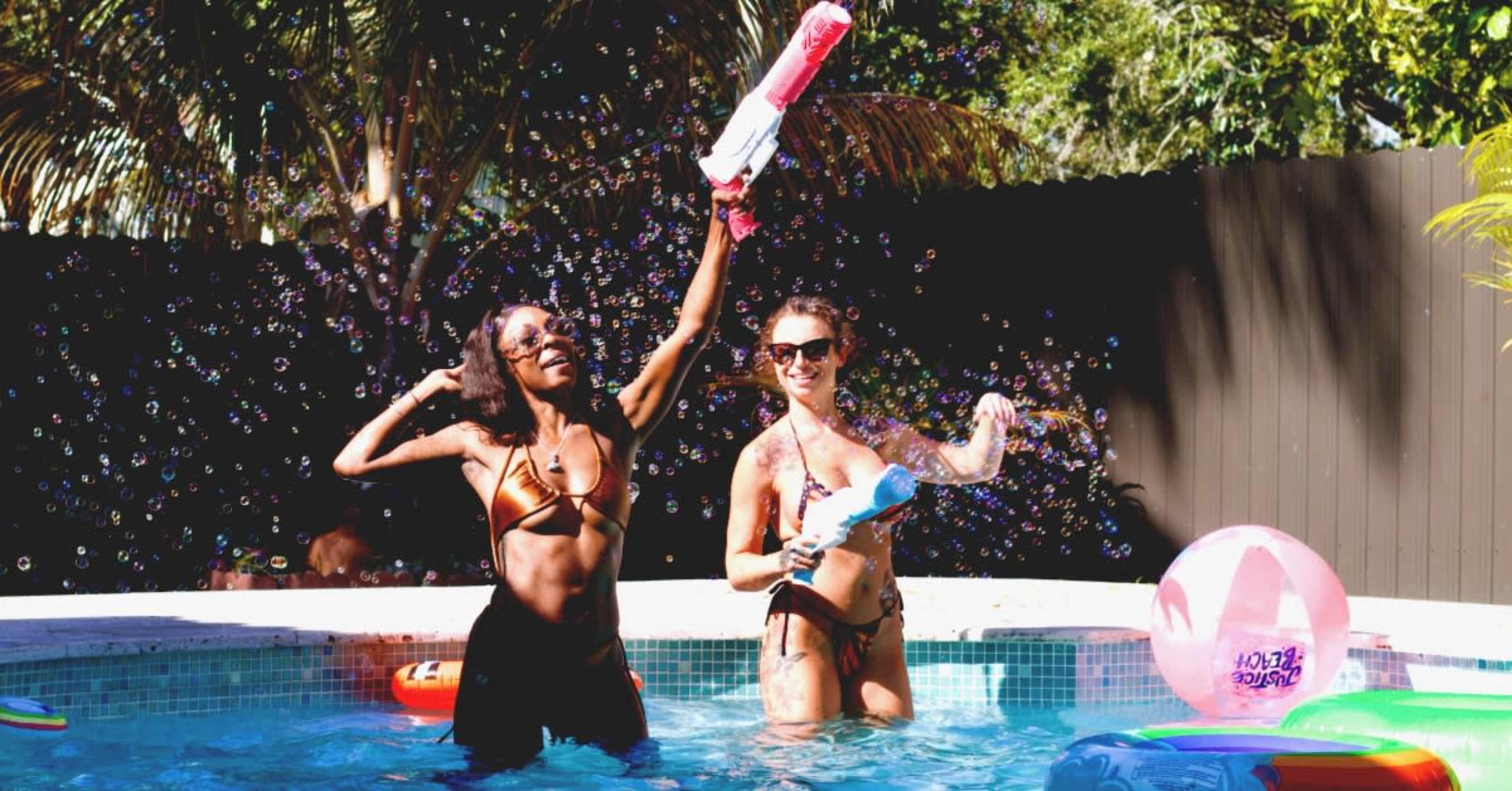 Two women play in a pool, holding water guns and surrounded by bubbles.