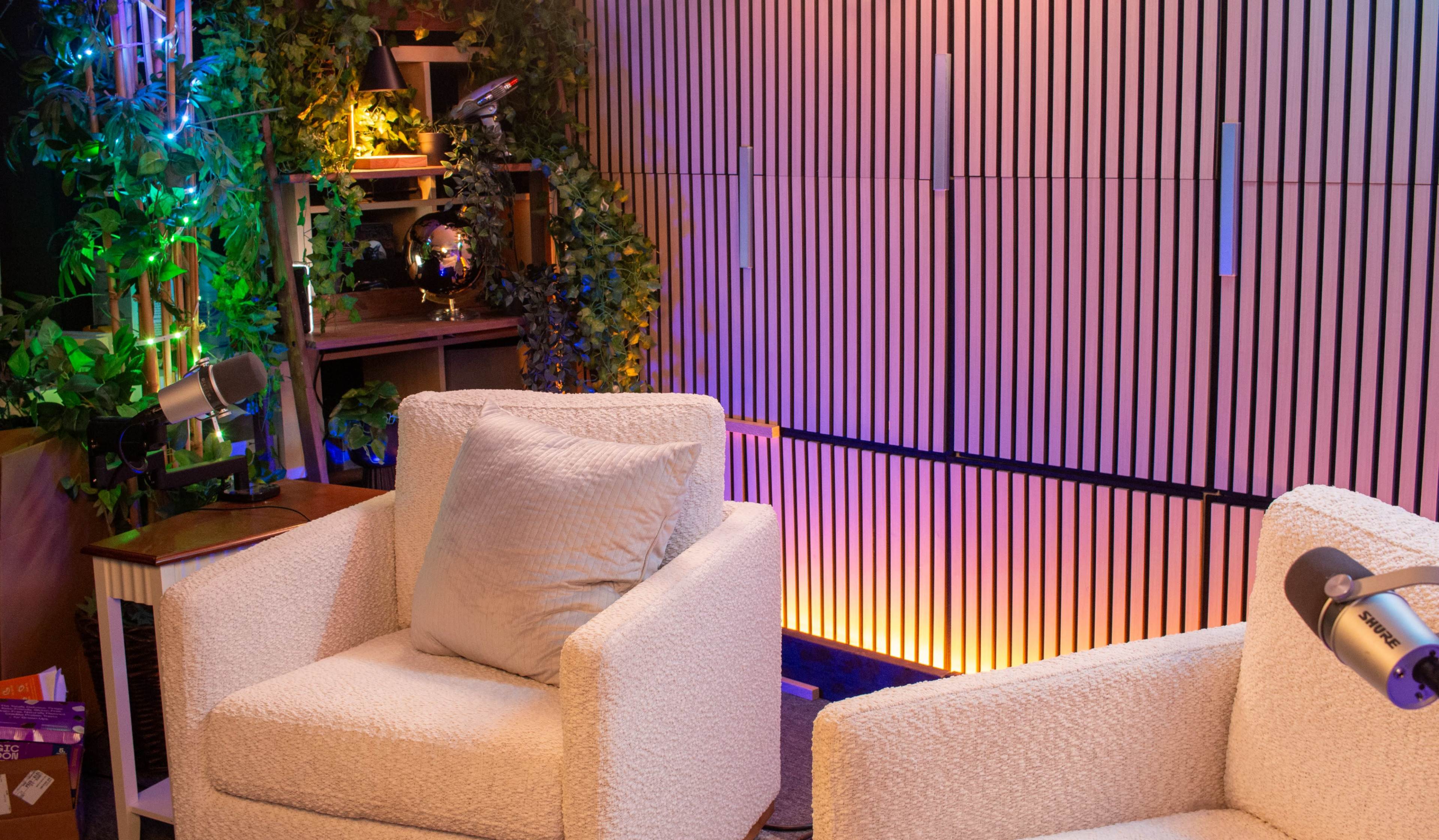 The image shows a cozy seating area with two white armchairs, surrounded by green plants and a striped wall featuring ambient lighting.