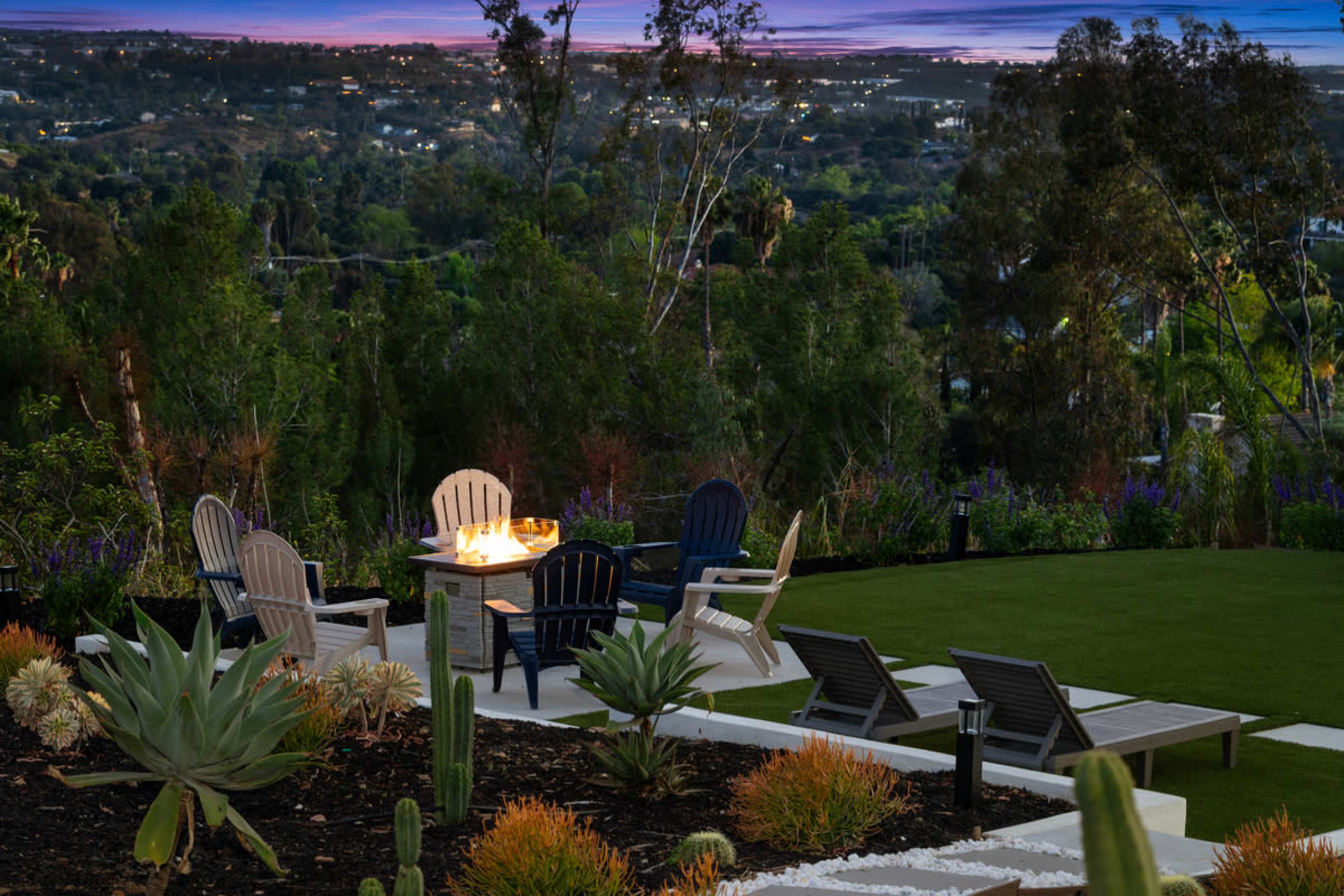 The image shows a backyard seating area with four chairs and a table surrounded by plants, overlooking a scenic landscape at dusk.