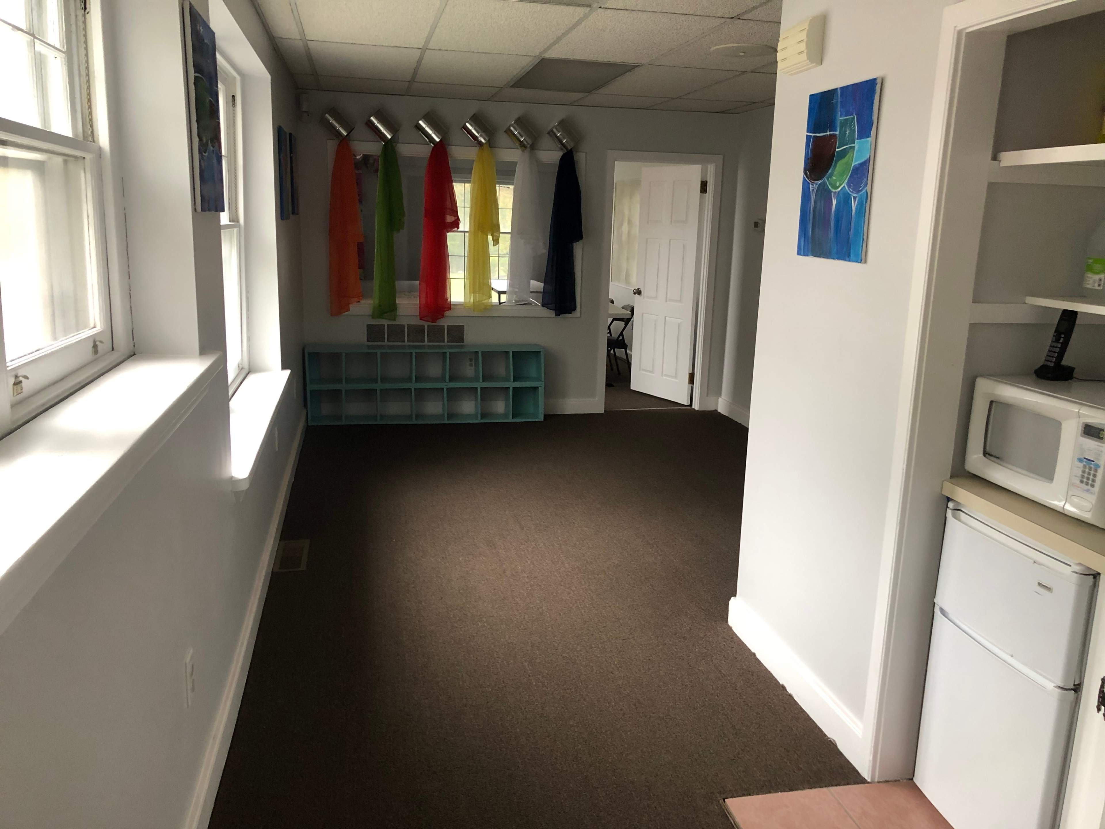 An interior room with a carpeted floor, white walls, a row of colored garments hanging from a rack, and a kitchenette area featuring a refrigerator and a microwave.