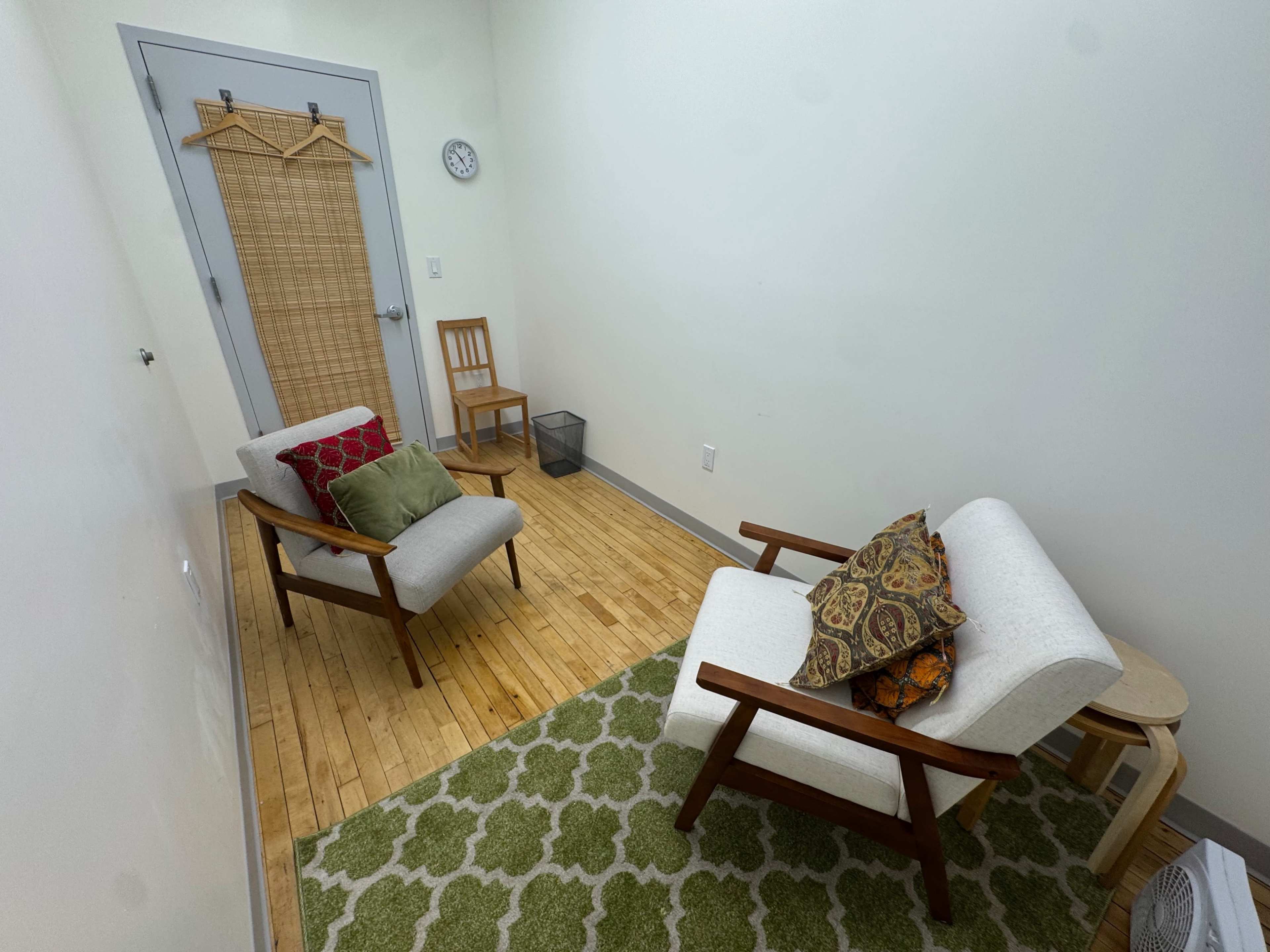The image shows a small, minimalist room with two mid-century modern chairs, a wooden side table, a small bookshelf, and a wall clock.