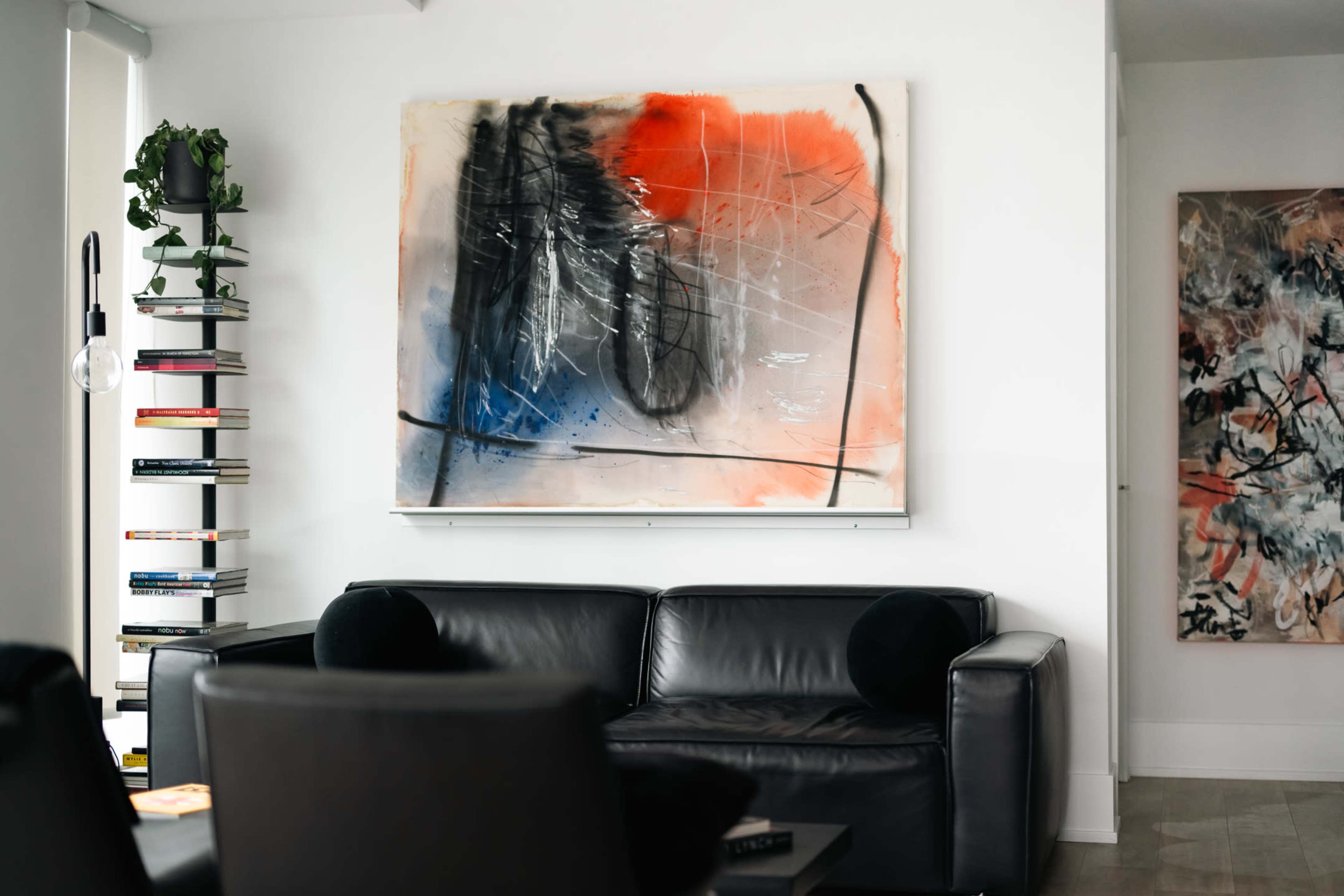 A black leather sofa sits in a modern room adorned with abstract artwork on the wall and a tall bookshelf filled with books.