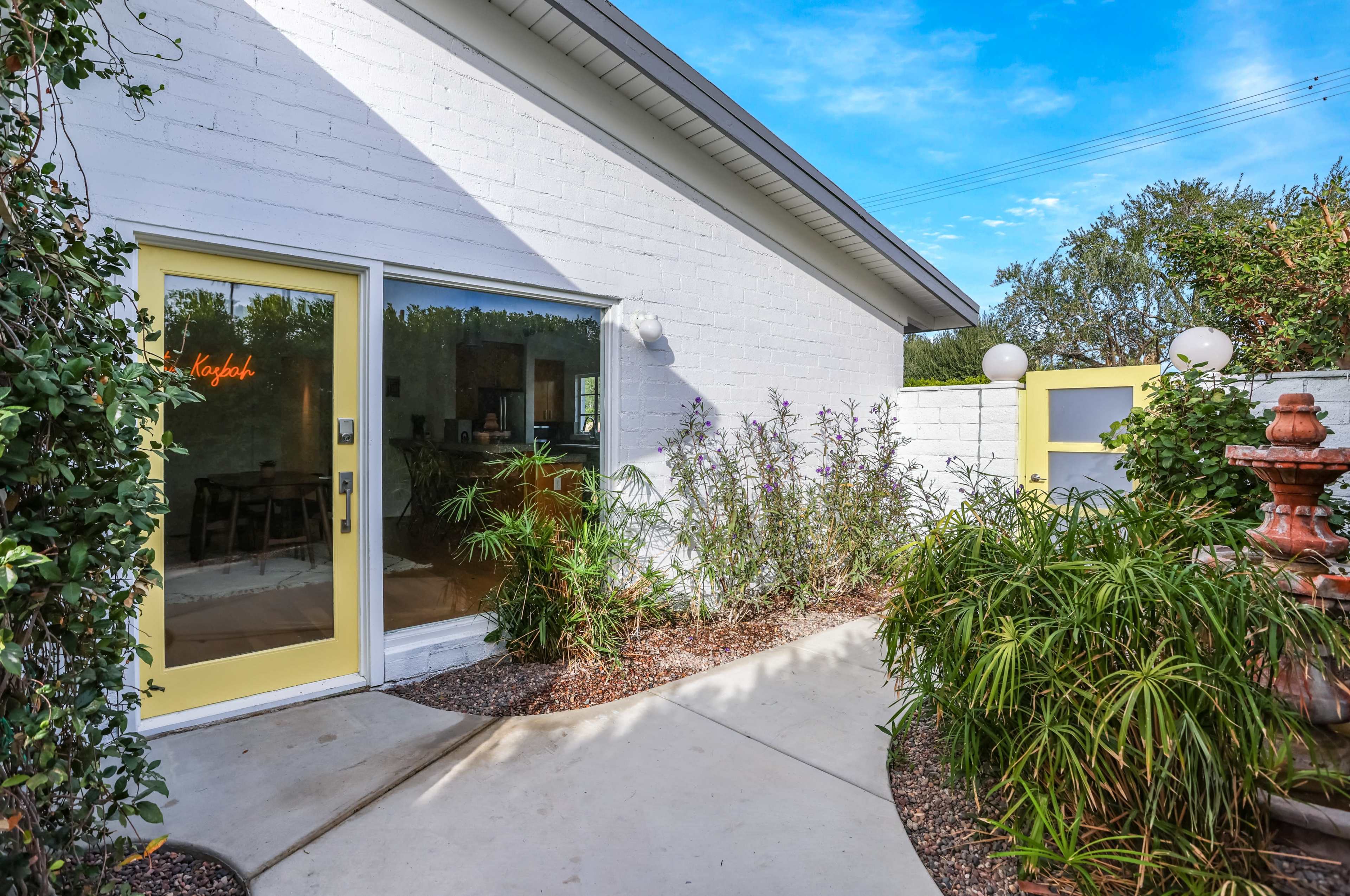 A bright yellow door leads into a white brick building, surrounded by greenery and a pathway.