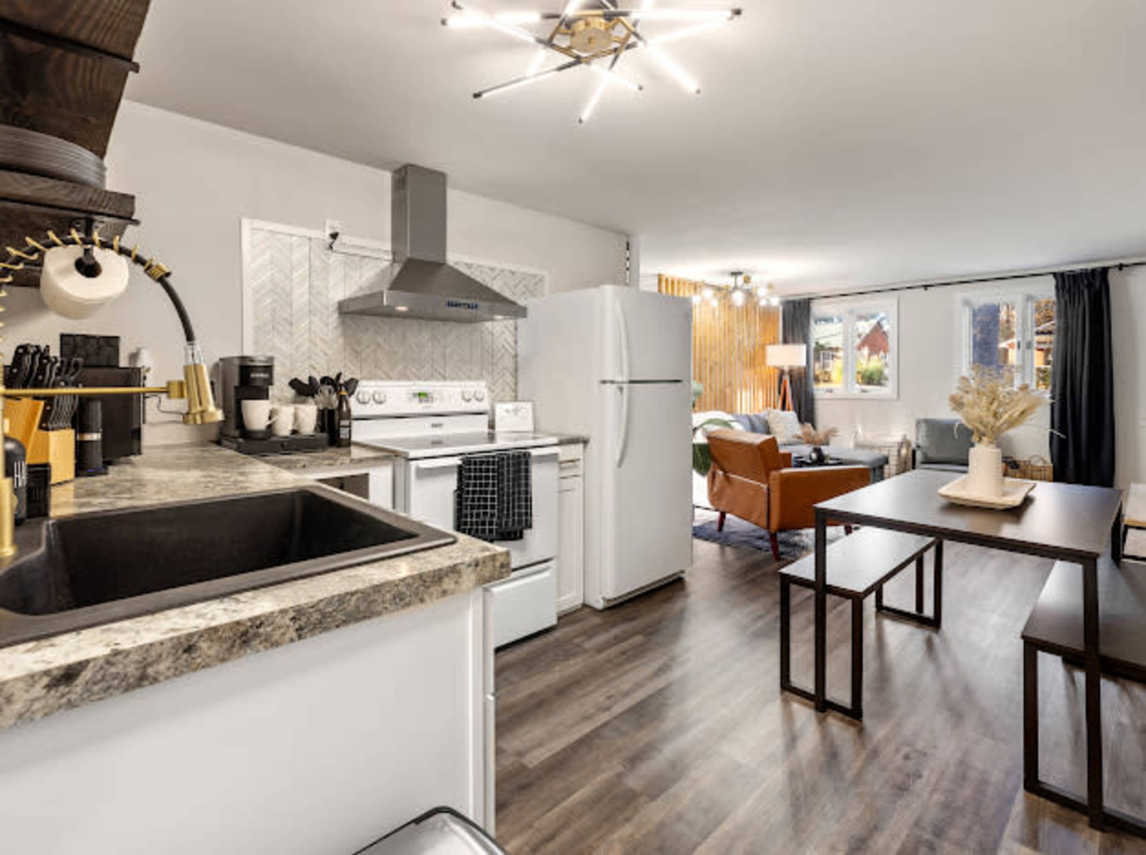 A modern kitchen features white appliances, a granite countertop, and a dining area with a table and chairs, leading to a cozy living room with large windows.