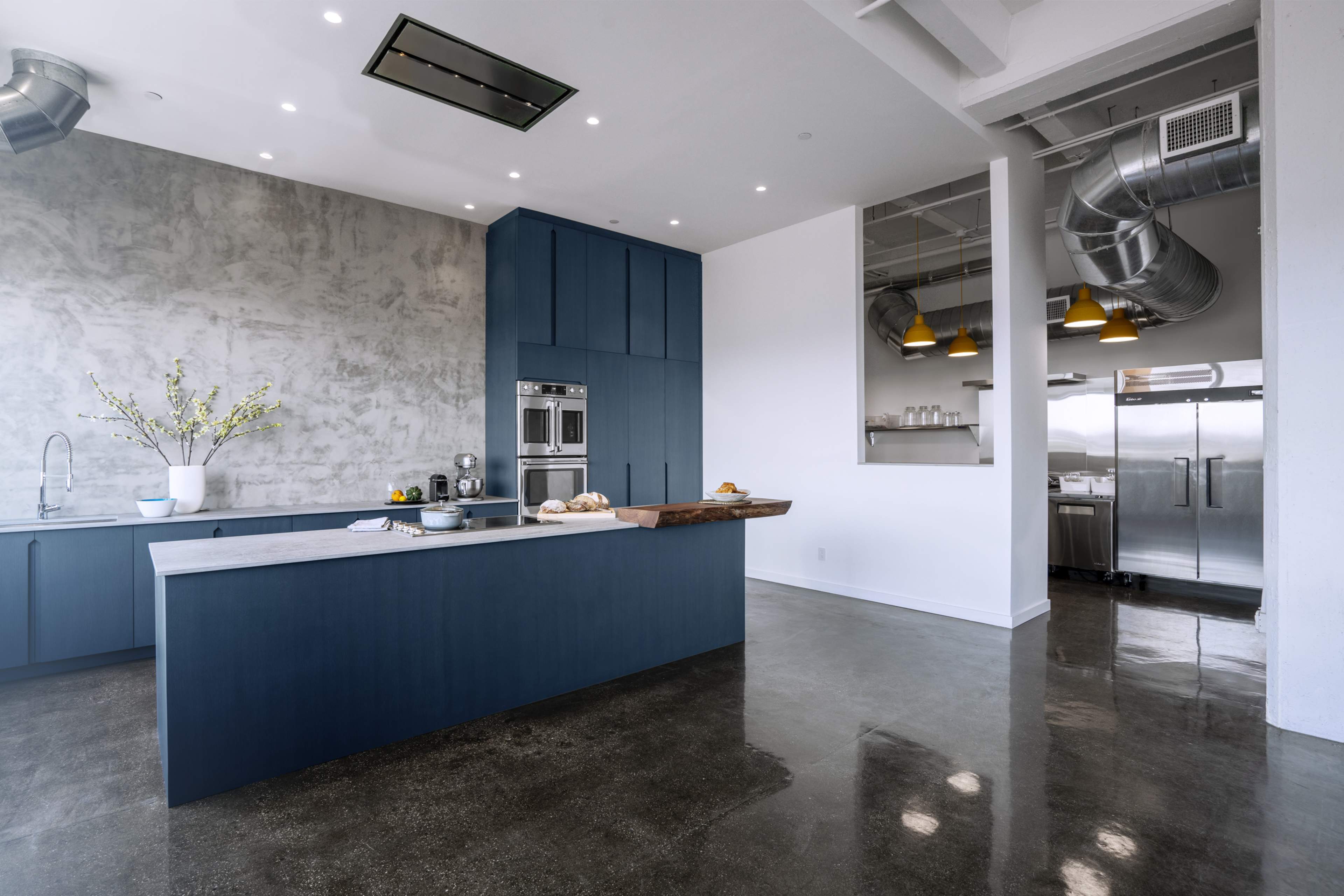 A modern kitchen features blue cabinetry, a large gray island with a wooden cutting board, and stainless steel appliances against a concrete wall.