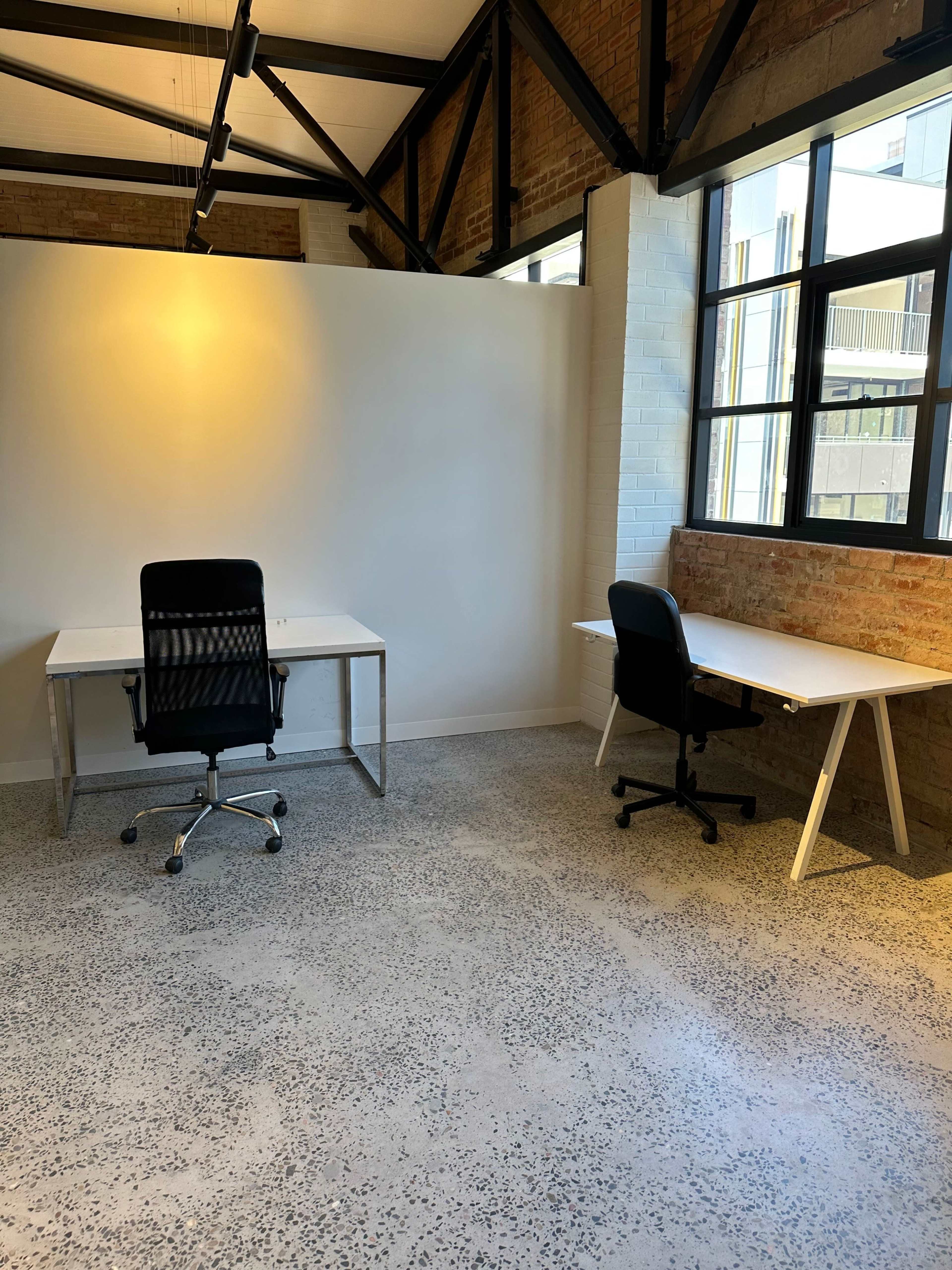 An office space with two empty desks and chairs on a polished concrete floor, separated by a white wall.