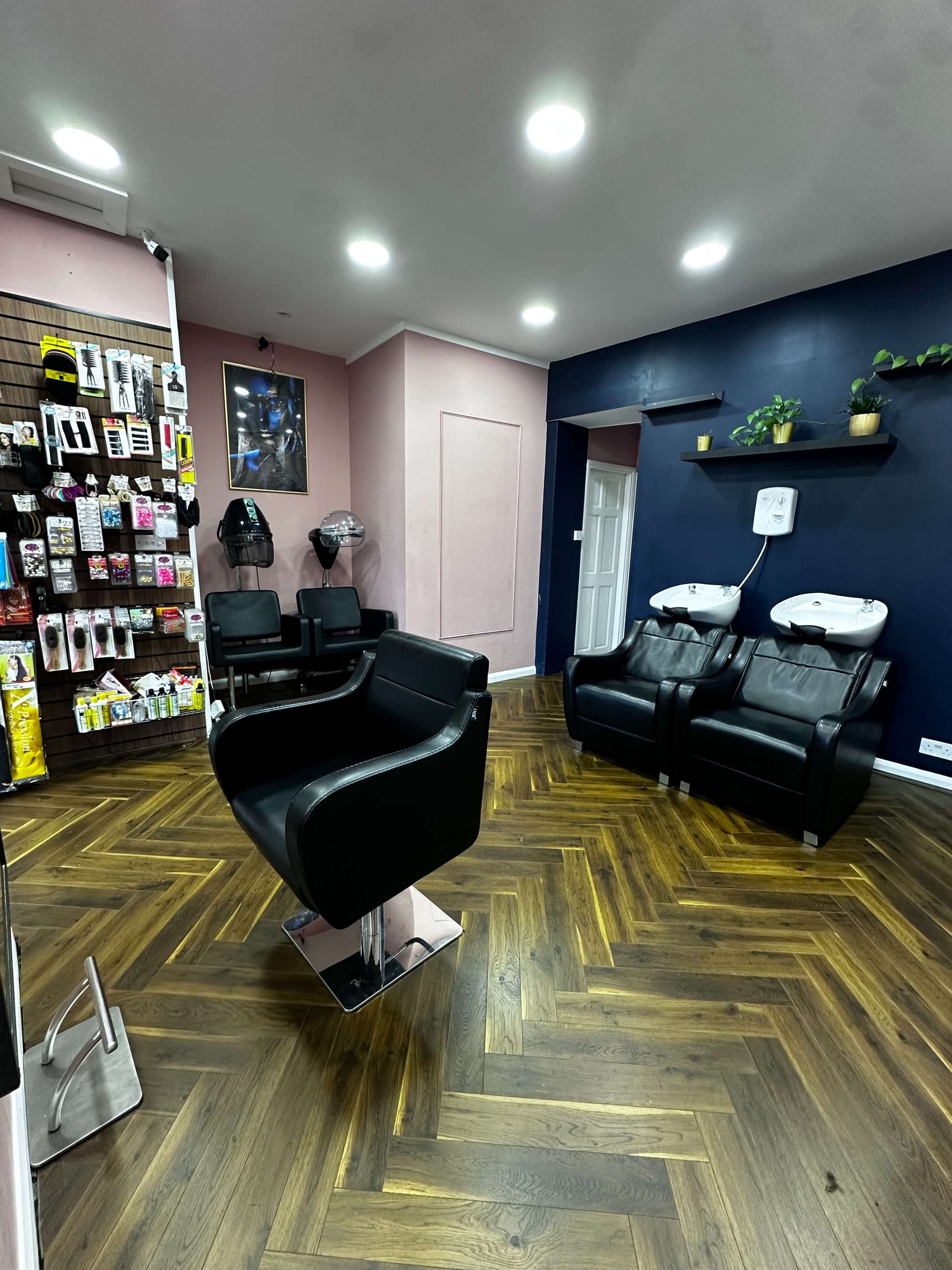 The image shows a modern hair salon featuring black styling chairs, two shampoo stations, a product display wall, and a hardwood floor arranged in a herringbone pattern.