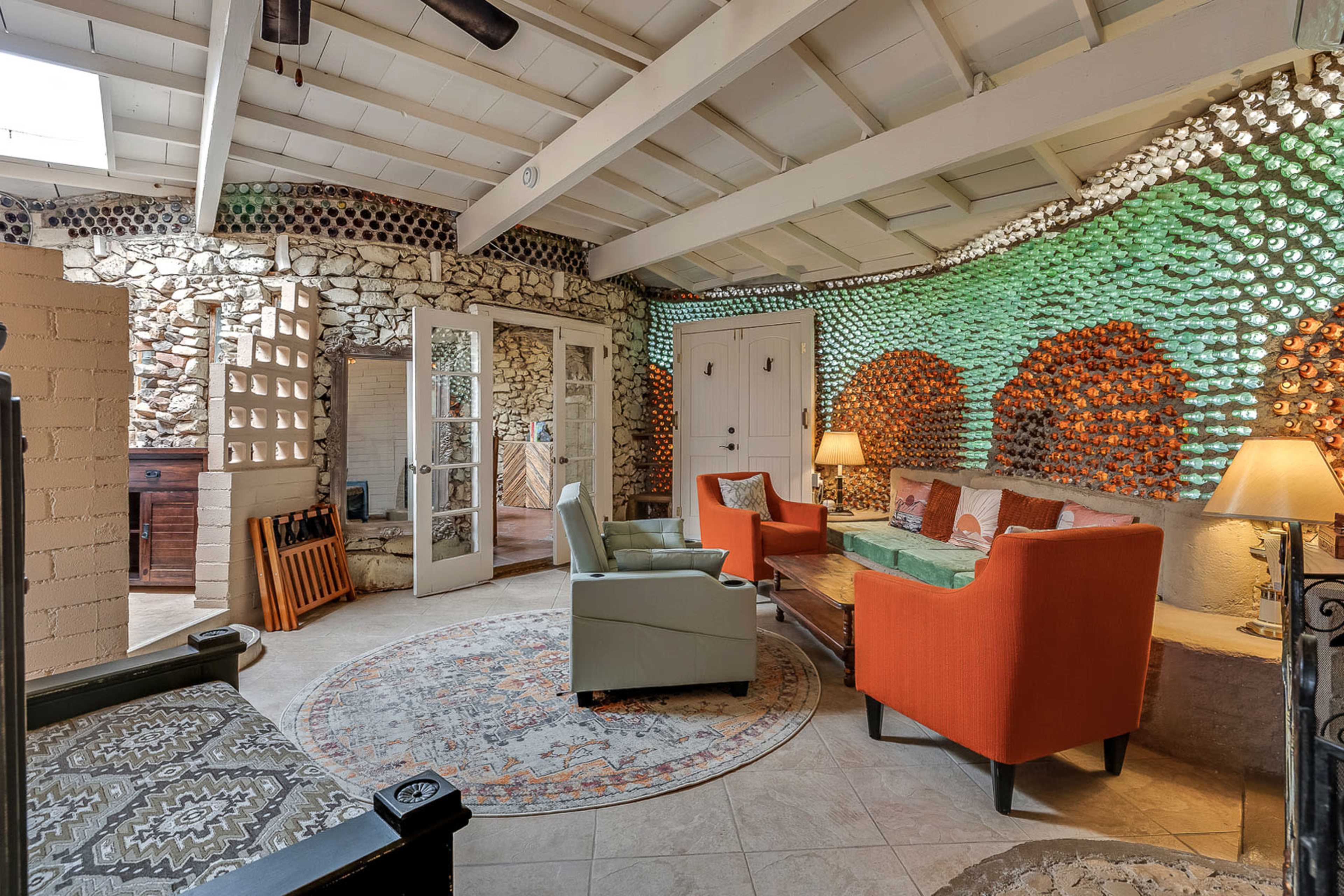 The room features a cozy seating area with orange chairs, a wooden table, and a unique wall decorated with colorful stones and bottles.